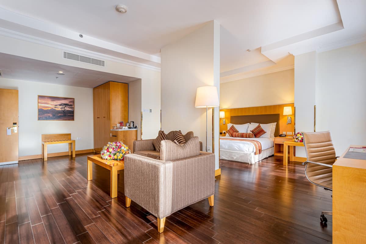 A hotel room with a sofa, a small table with a vase, a bed with pillows, a nightstand with a lamp, and a wooden chair.