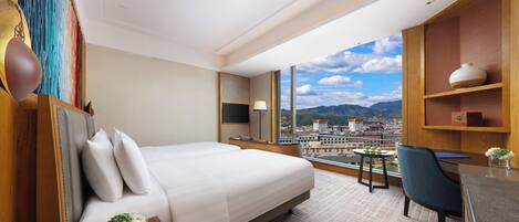 A hotel room with a large bed, a desk, a chair, a TV, and a view of a cityscape.