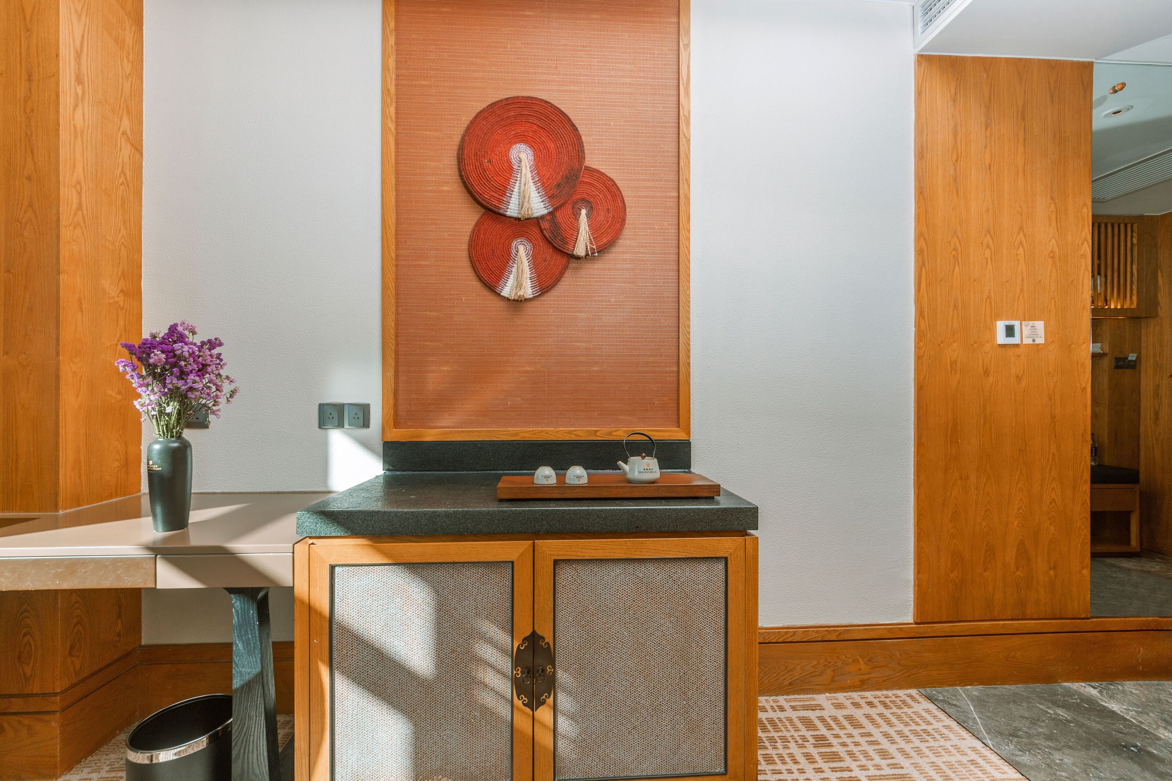 Un mueble de madera con puerta de mimbre, un adorno de pared y una pequeña estantería con una tetera y un jarrón de flores.