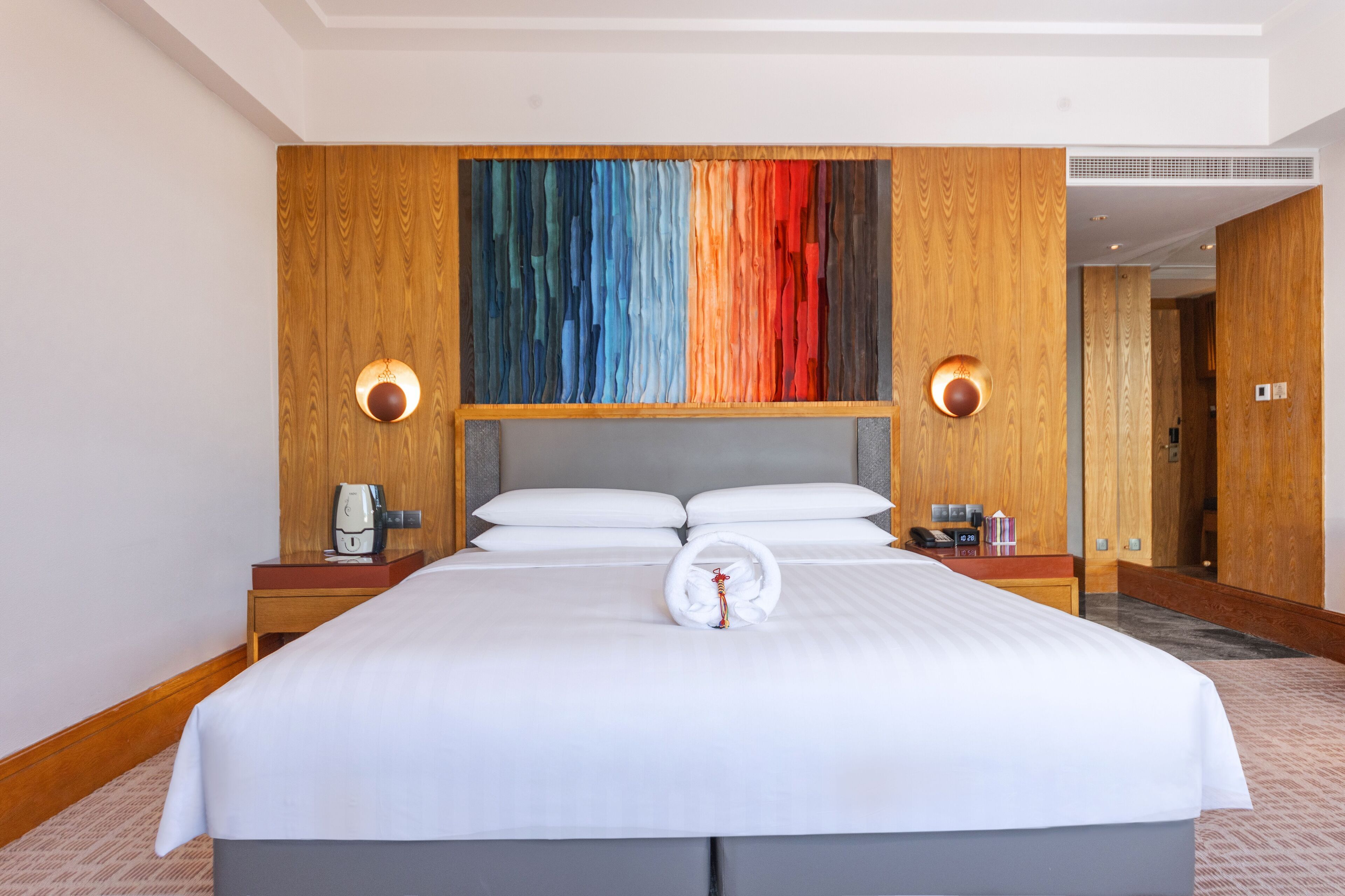 Camera d'albergo con un letto grande, una testiera in legno, un'opera d'arte astratta e colorata sopra il letto, due comodini con lampade e un letto rifatto con un asciugamano e una sveglia.