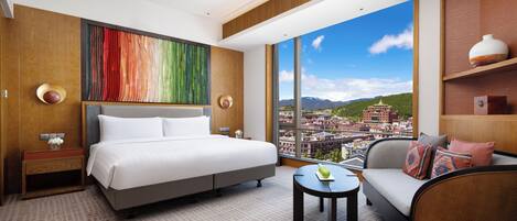 A hotel room with a large bed, a sofa, a small table, and a view of a cityscape through large windows.