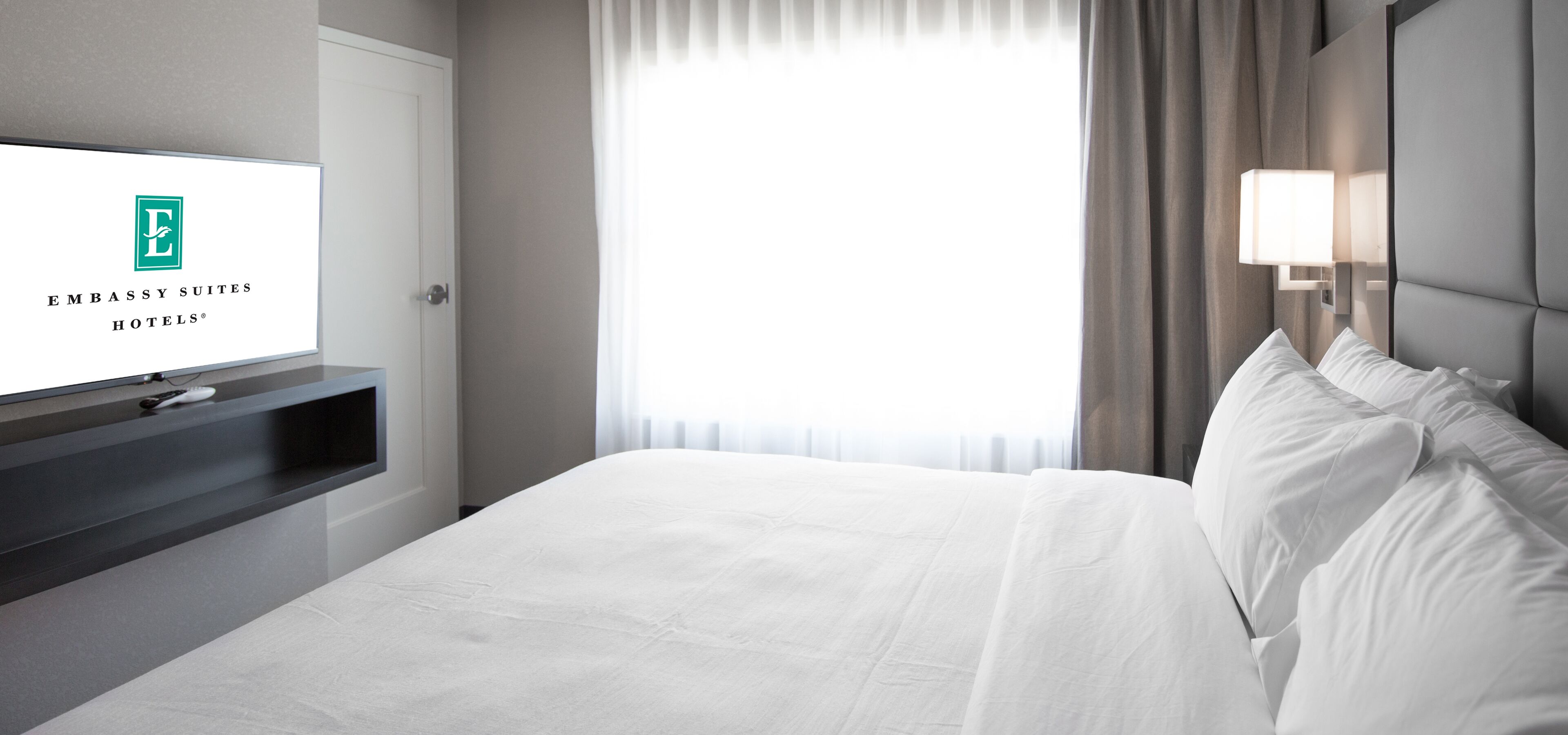 A hotel room with a bed, a TV on a stand, a window with curtains, and a lamp on the wall.