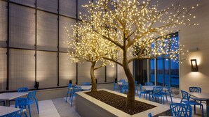 A modern indoor dining area with a tree illuminated by lights, blue chairs, and tables. - Embassy Suites by Hilton Kansas City Olathe (Olathe)