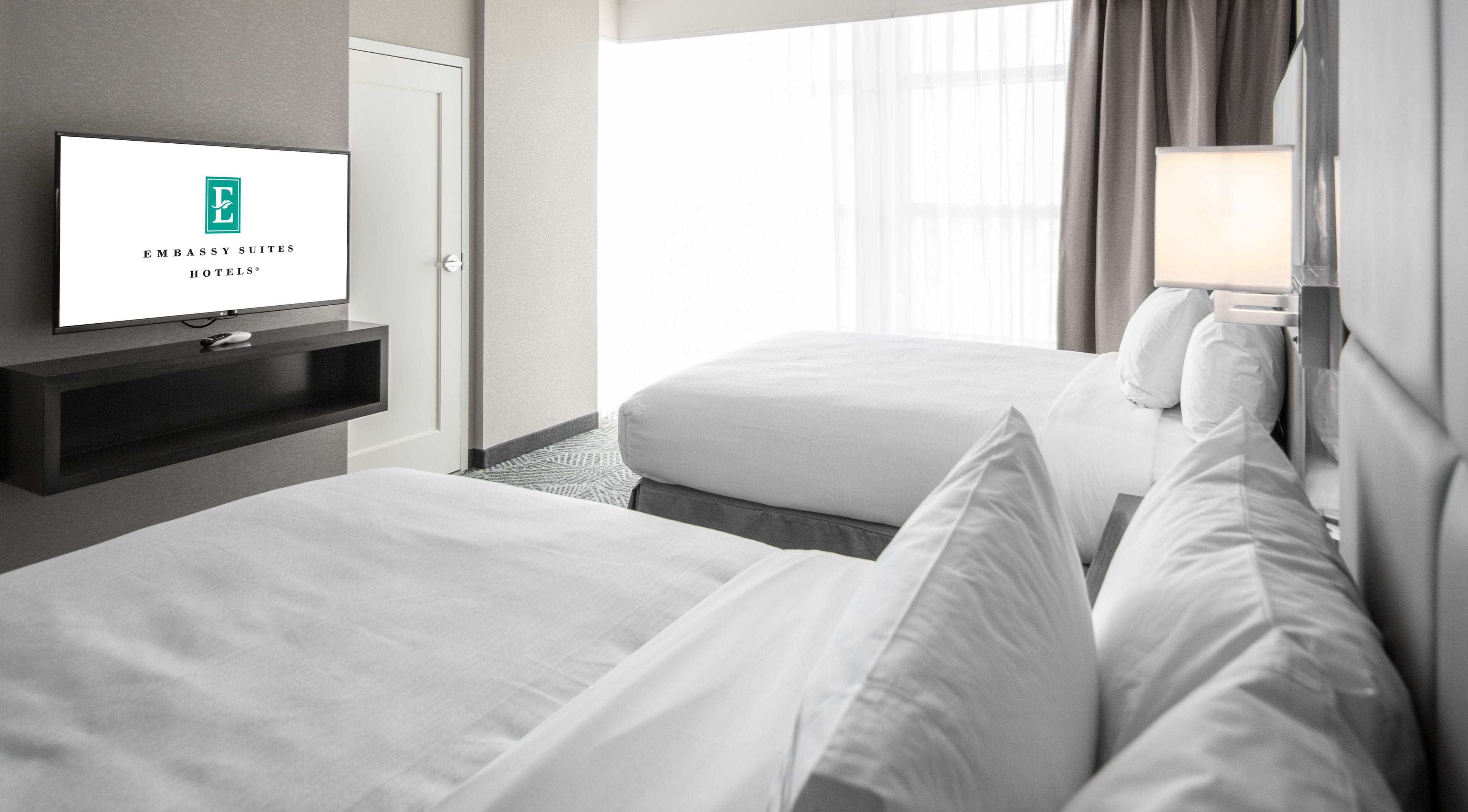 A hotel room with two beds, a TV on a stand, a window with curtains, and a lamp.