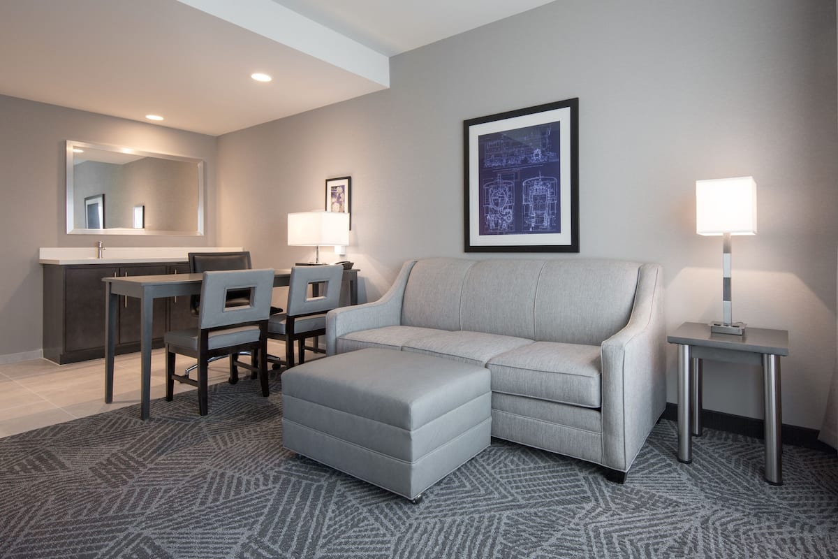 Une chambre d’hôtel moderne avec un canapé gris, une table à manger avec des chaises, un petit bureau et un tableau encadré au mur.