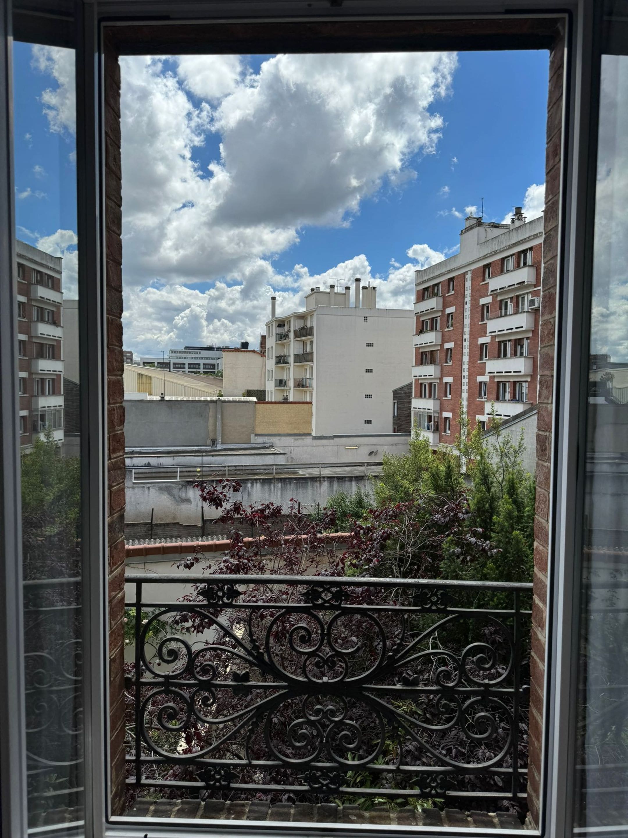 A view from a window with a metal balcony railing, overlooking a cityscape with multi-story buildings and a clear sky with clouds.
