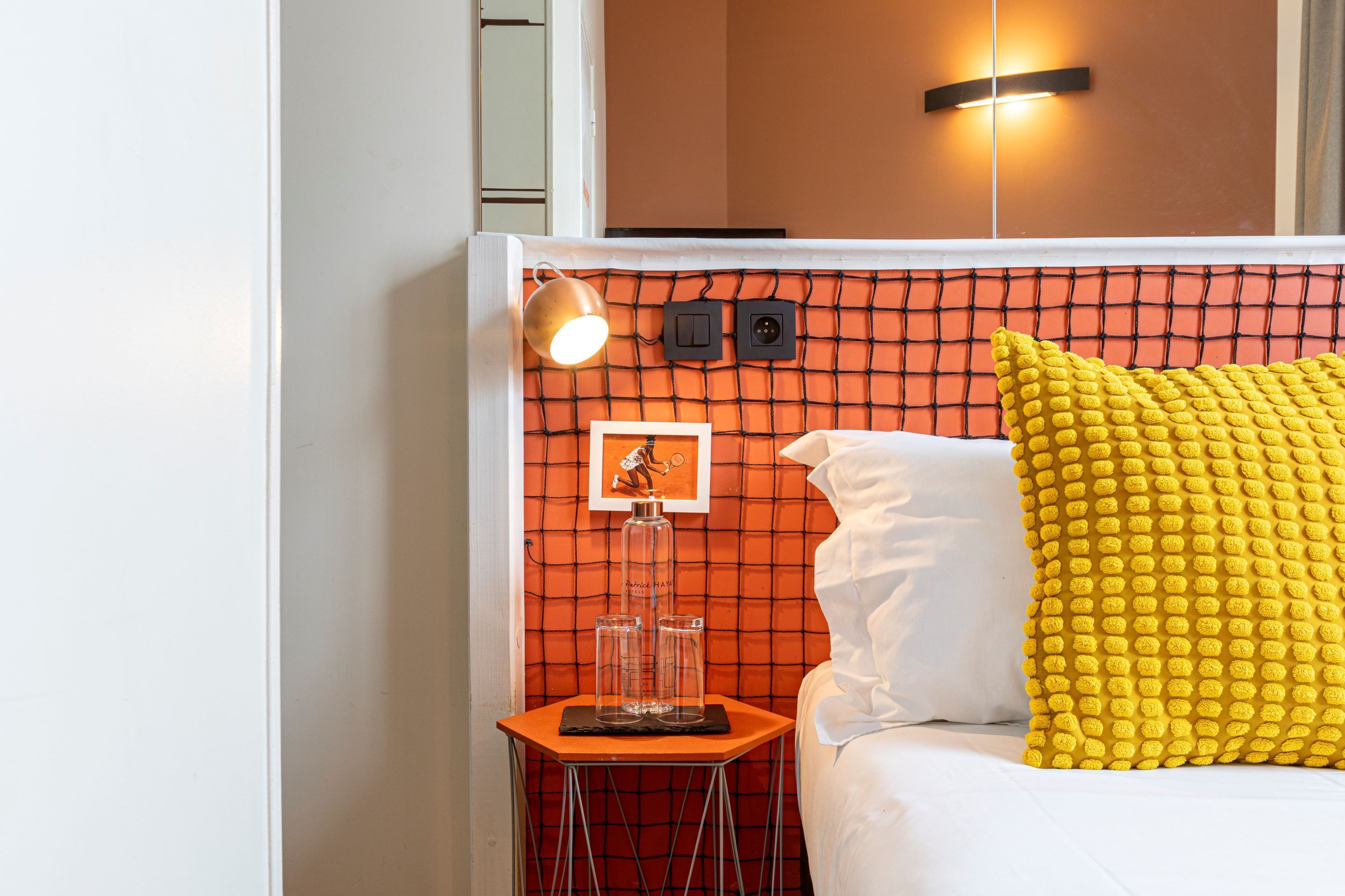 A bedroom with a bed, a bedside table with a lamp, a wall-mounted light, and a picture frame.