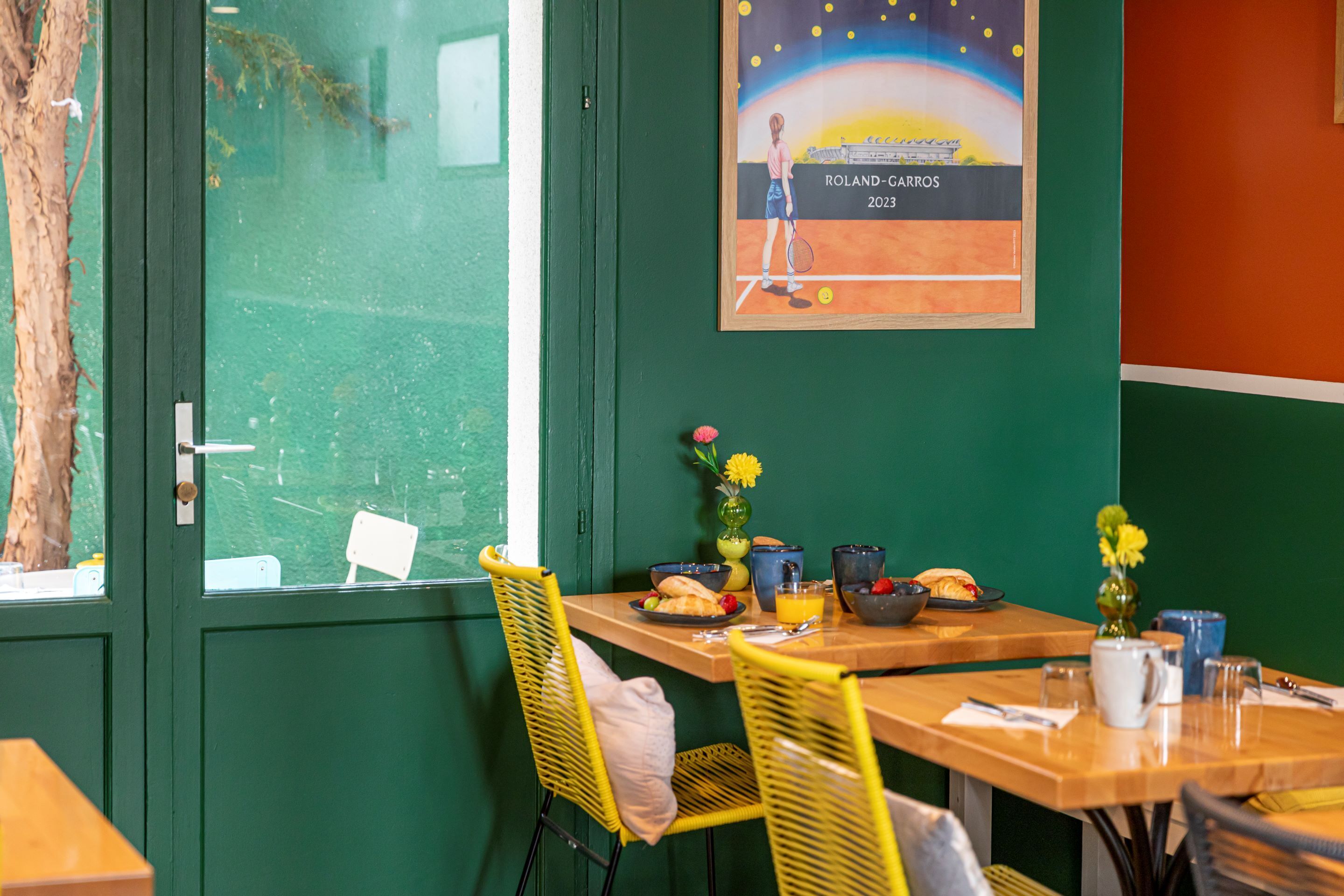 Een eethoek met een tafel gedekt voor twee personen, met een poster met tennisthema, gele stoelen en een vaas met bloemen.