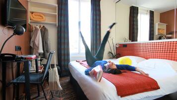 A hotel room with a bed, a desk with a lamp, a chair, a shelf with towels, and a window with curtains.