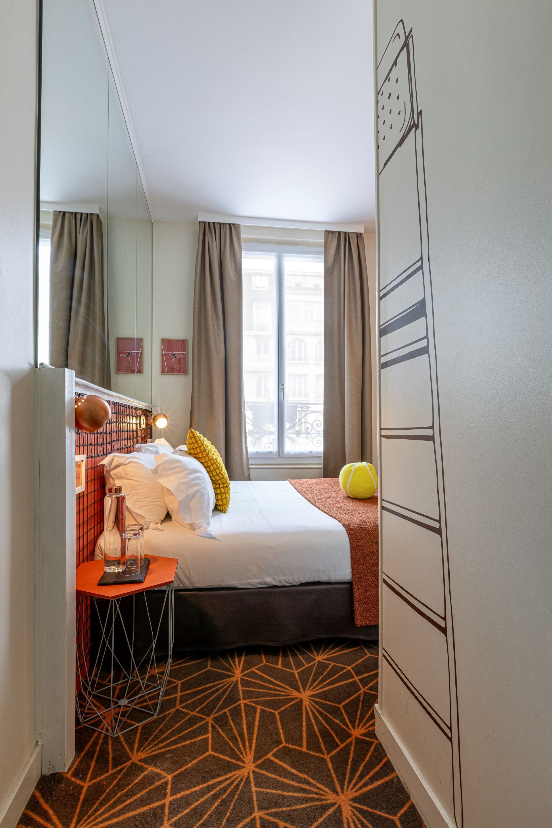 Una habitación de hotel con una cama, una mesita con una lámpara, una ventana con cortinas y una alfombra de estampado geométrico.