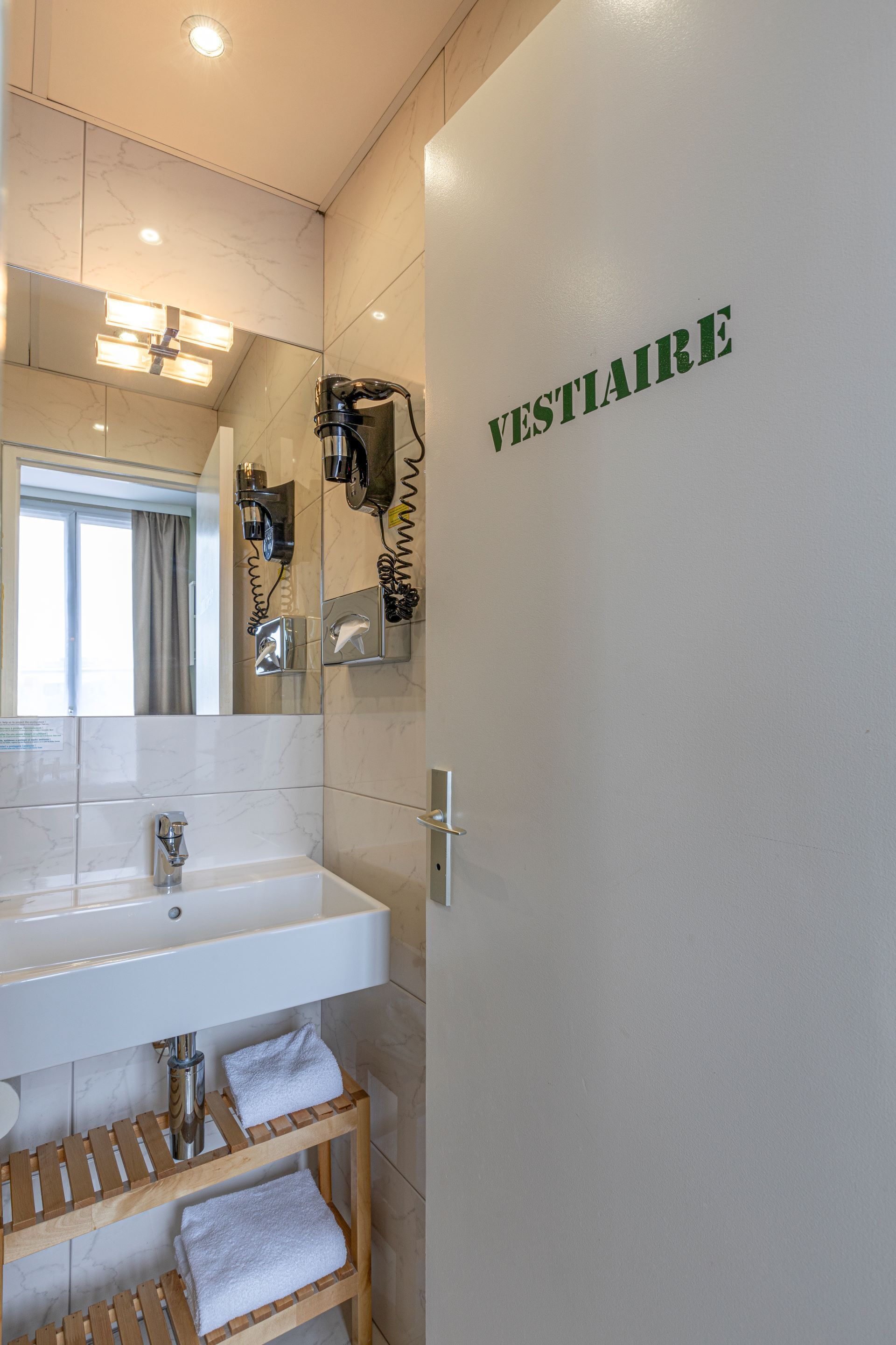 A bathroom with a white sink, mirror, and towel racks. The word "VESTIAIRE" is visible on the wall.