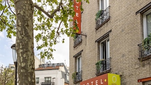 A hotel with a red and yellow sign, brick facade, and balconies with plants. - Hotel Olympic Paris Boulogne by Patrick Hayat (Boulogne-Billancourt)