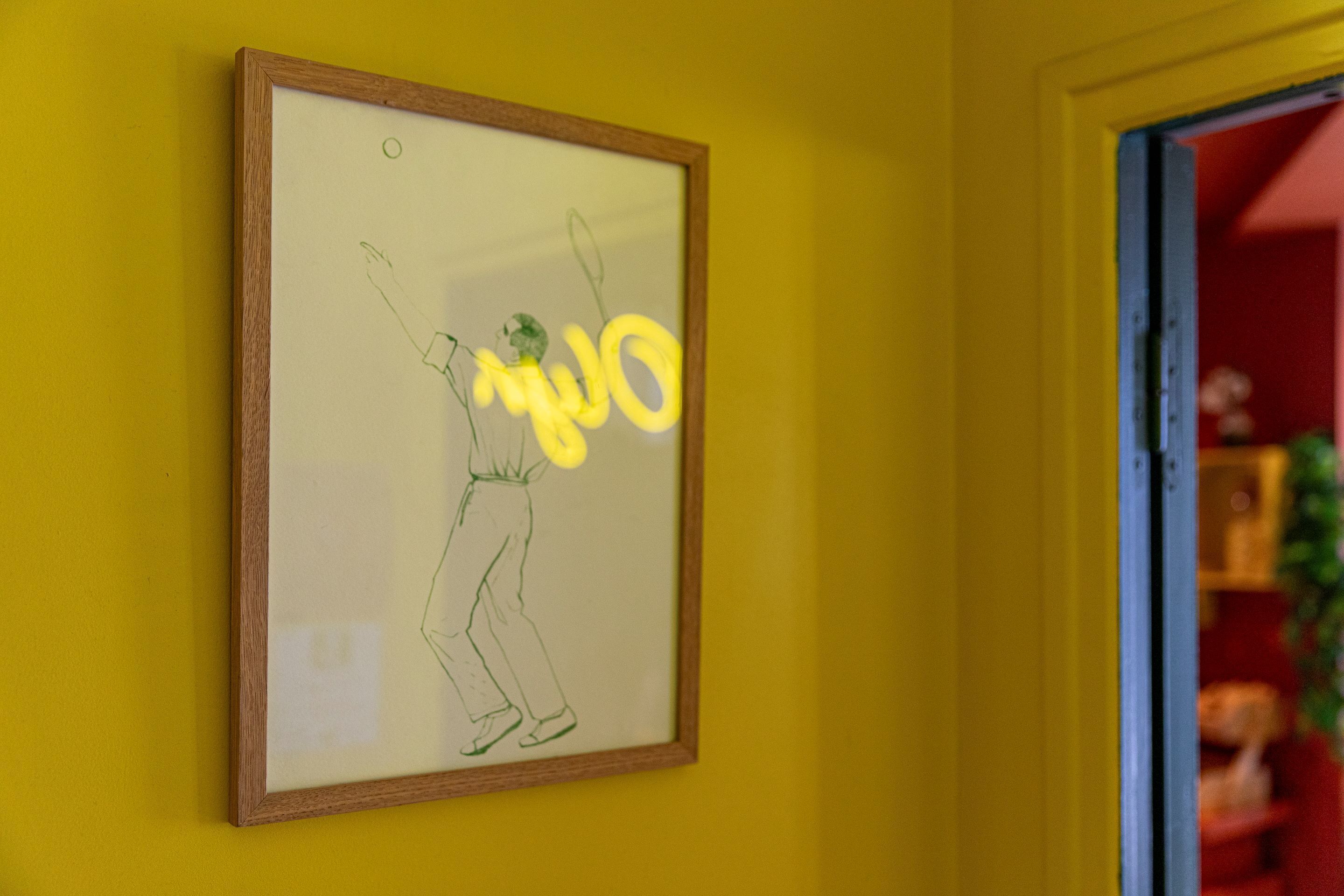 A framed drawing of a person playing tennis on a yellow wall.