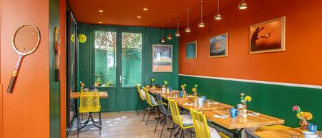 A dining area with wooden tables and yellow chairs, green walls, and tennis rackets on the left.