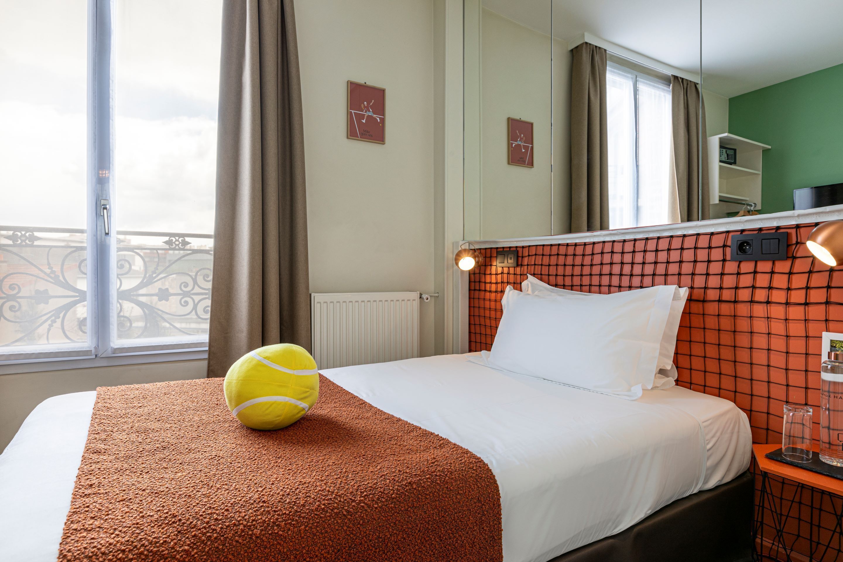 A hotel room with a bed, a tennis ball on the bed, a nightstand with a glass of water, and a view of the outside balcony.