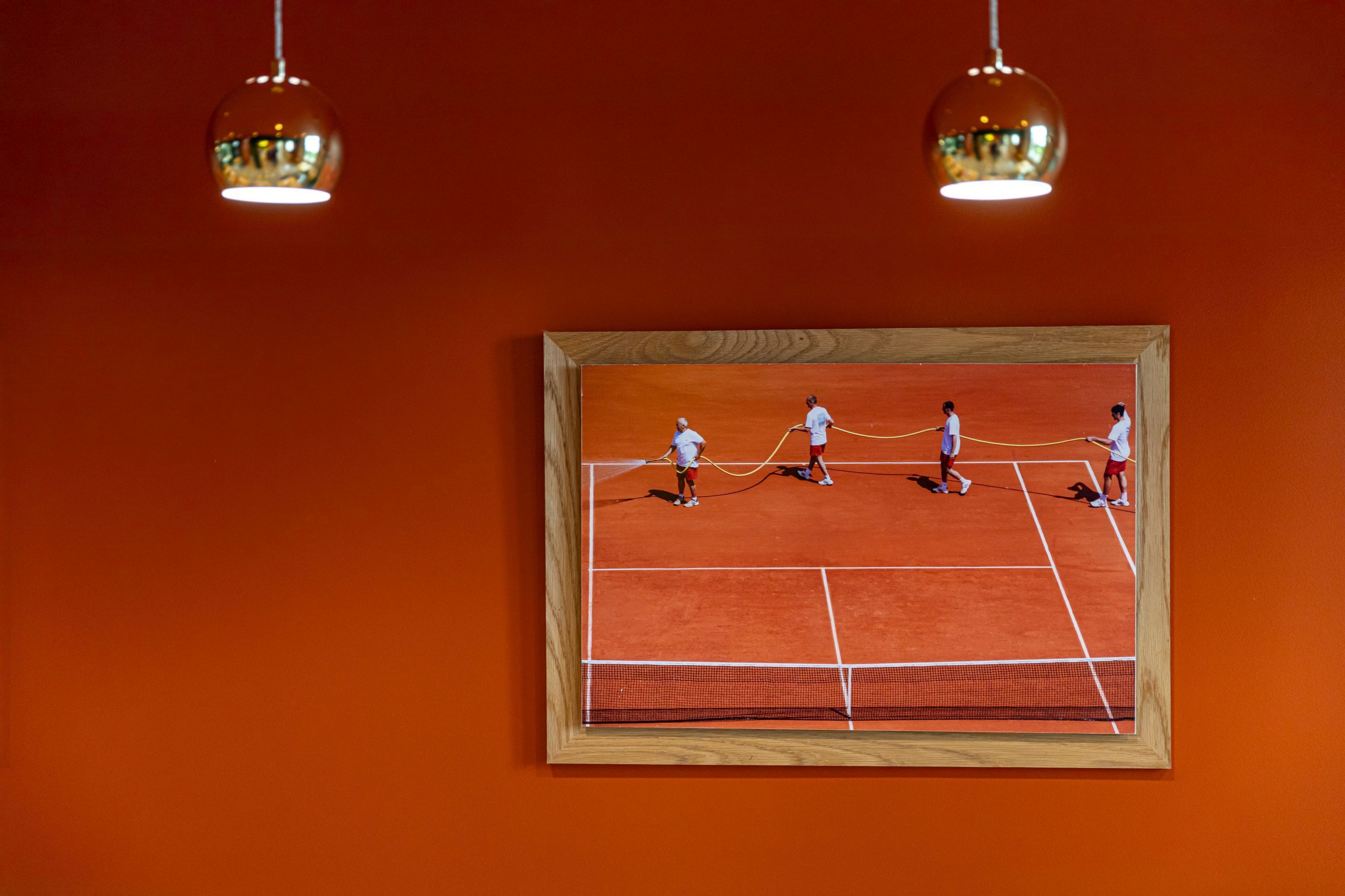 A framed tennis court painting on an orange wall with two hanging pendant lights.