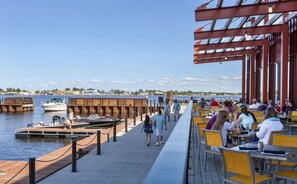 A waterfront restaurant with outdoor seating overlooking the water, a dock with boats, and a clear view of the city skyline in the background.