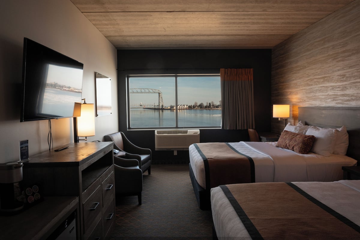 Une chambre d’hôtel avec un lit, un bureau avec une lampe, une chaise et une grande fenêtre donnant sur un pont et sur l’eau.