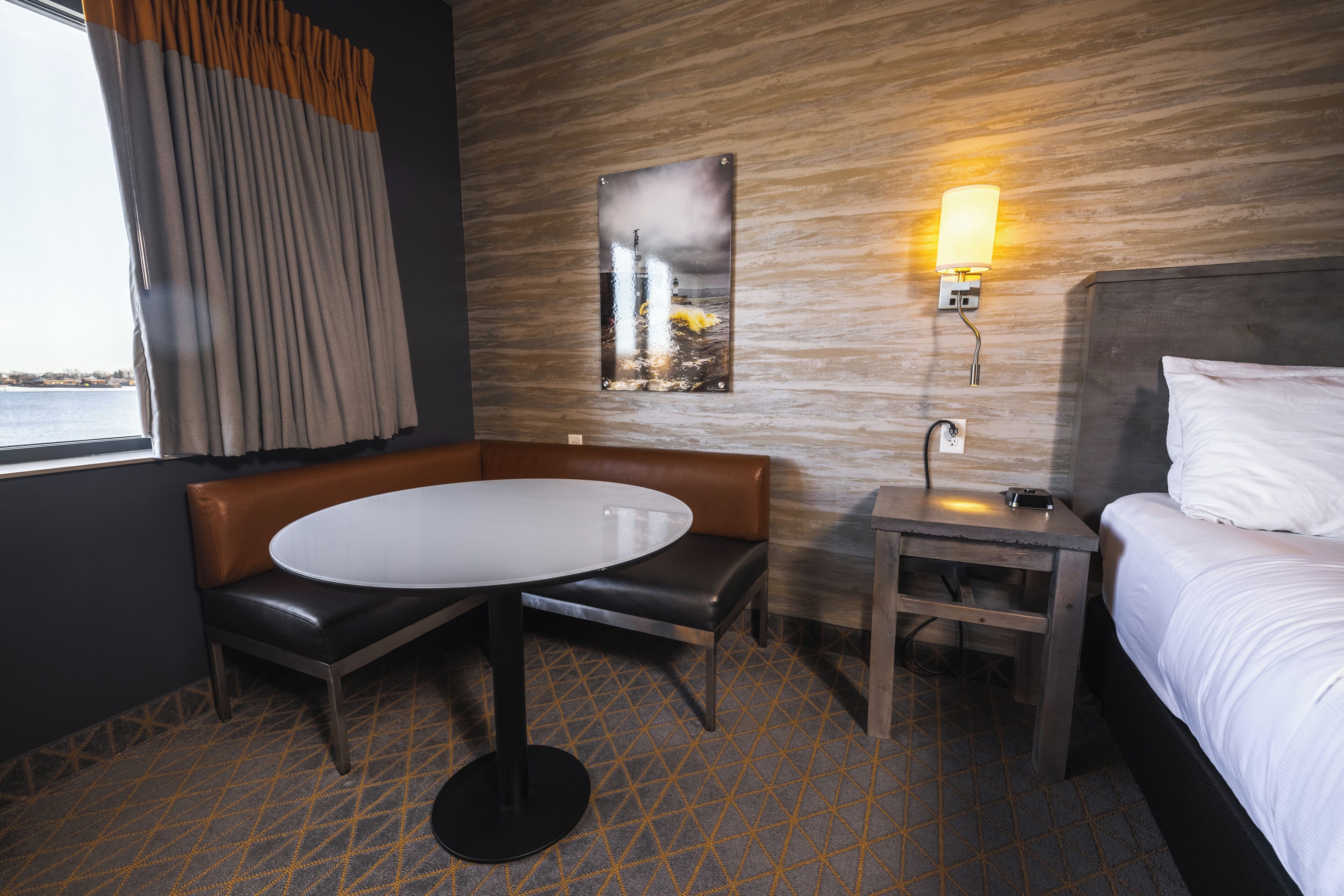 A hotel room with a bed, a small table, a bench, and a wall-mounted lamp.