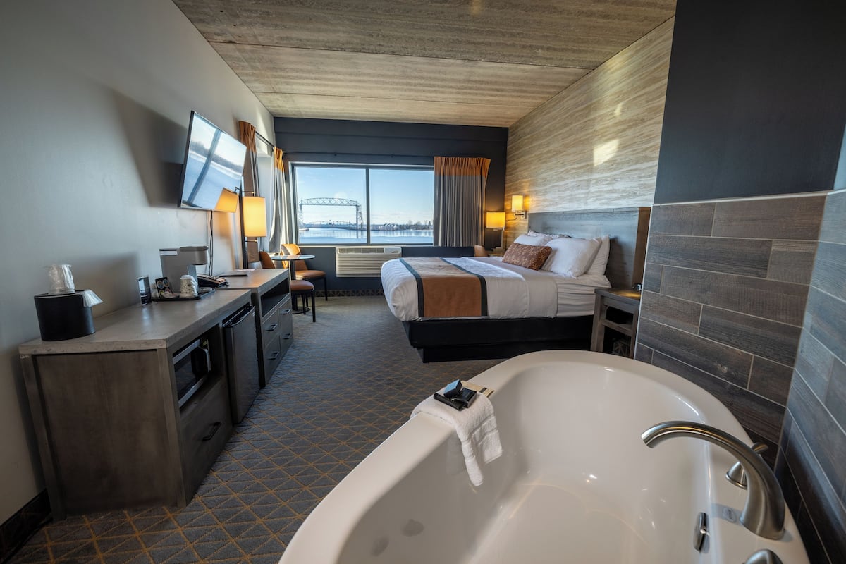 Une chambre d’hôtel avec un grand lit, un bureau avec un ordinateur, une lampe de bureau, une télévision, une fenêtre donnant sur un pont et une baignoire.