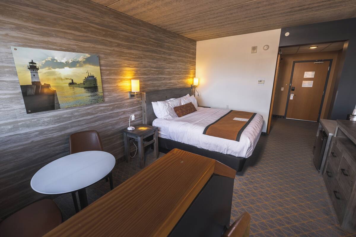 Une chambre d’hôtel avec un lit, des tables de chevet, une petite table et un tableau représentant un phare, accroché au mur.