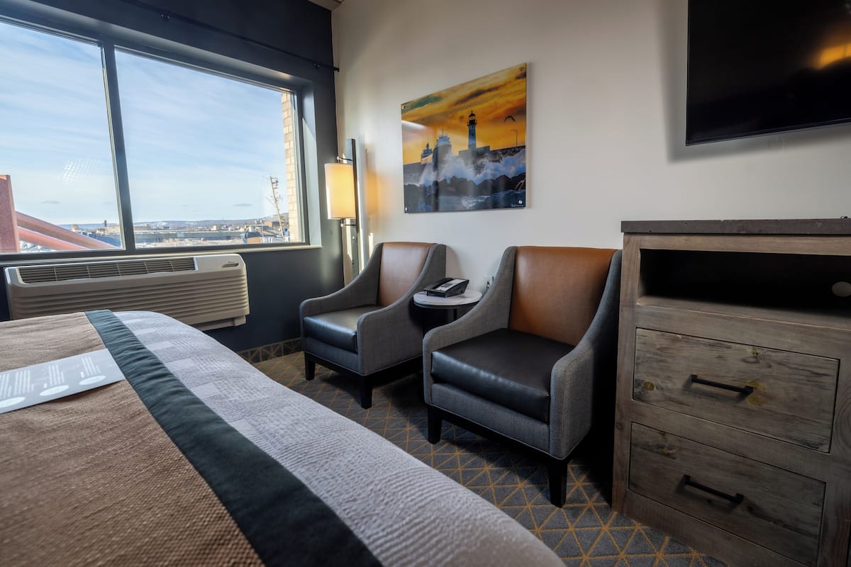 Une chambre d’hôtel comprenant un lit, deux fauteuils, une commode en bois, un téléviseur et un tableau représentant un paysage urbain.