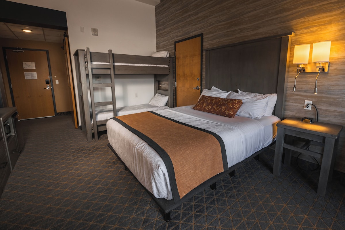 Une chambre d’hôtel avec un lit superposé, un lit simple, une table de chevet avec une lampe et un luminaire fixé au mur.