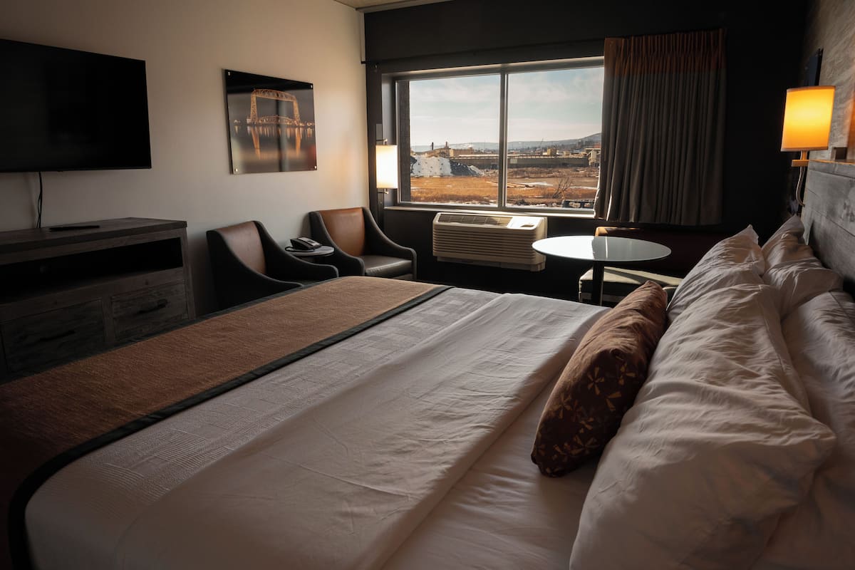 Une chambre d’hôtel avec un grand lit, un bureau, une chaise, une télévision et une fenêtre donnant sur un pont et un paysage.