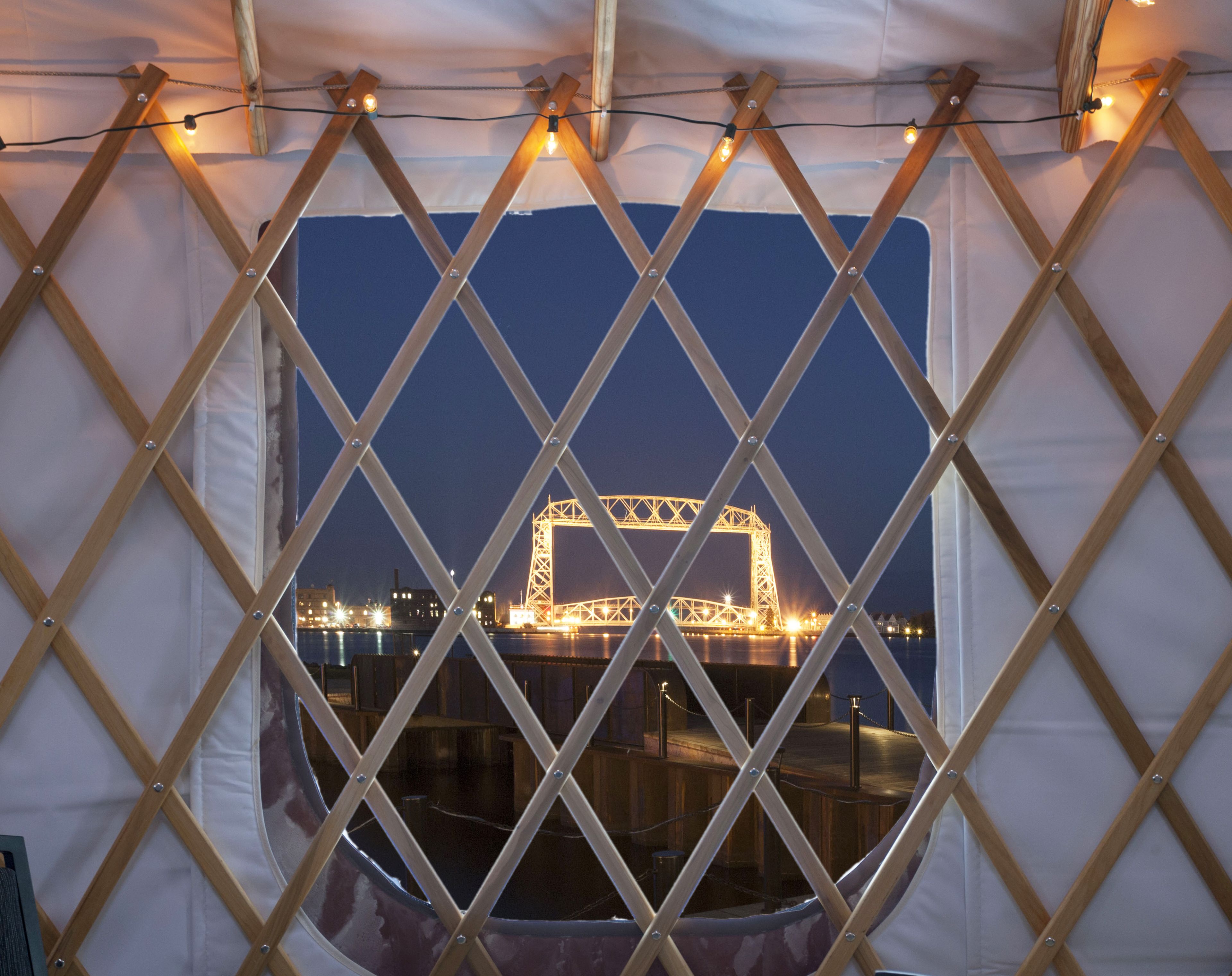 A view through a window with a wooden lattice frame, featuring a cityscape with illuminated structures at night.