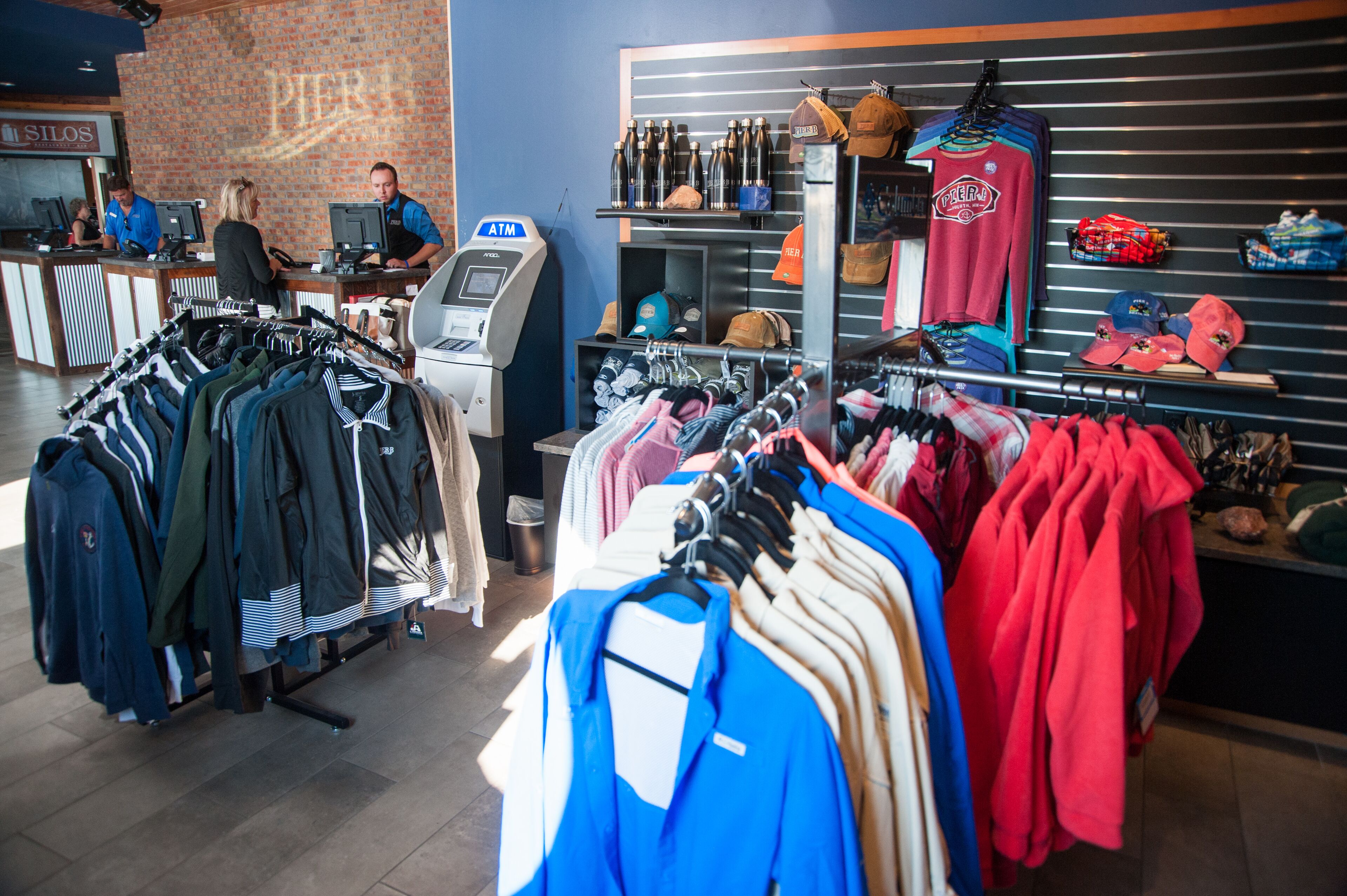 Un magasin de vêtements où l’on trouve des présentoirs remplis de vestes et de chemises, un distributeur automatique de billets et des étagères avec des chapeaux et du matériel de sport.