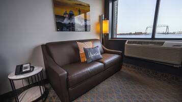 Habitación de hotel con un sofá de cuero marrón, una mesita auxiliar con un teléfono, una lámpara y un ventanal que ofrece vistas a un puente y al paisaje urbano.