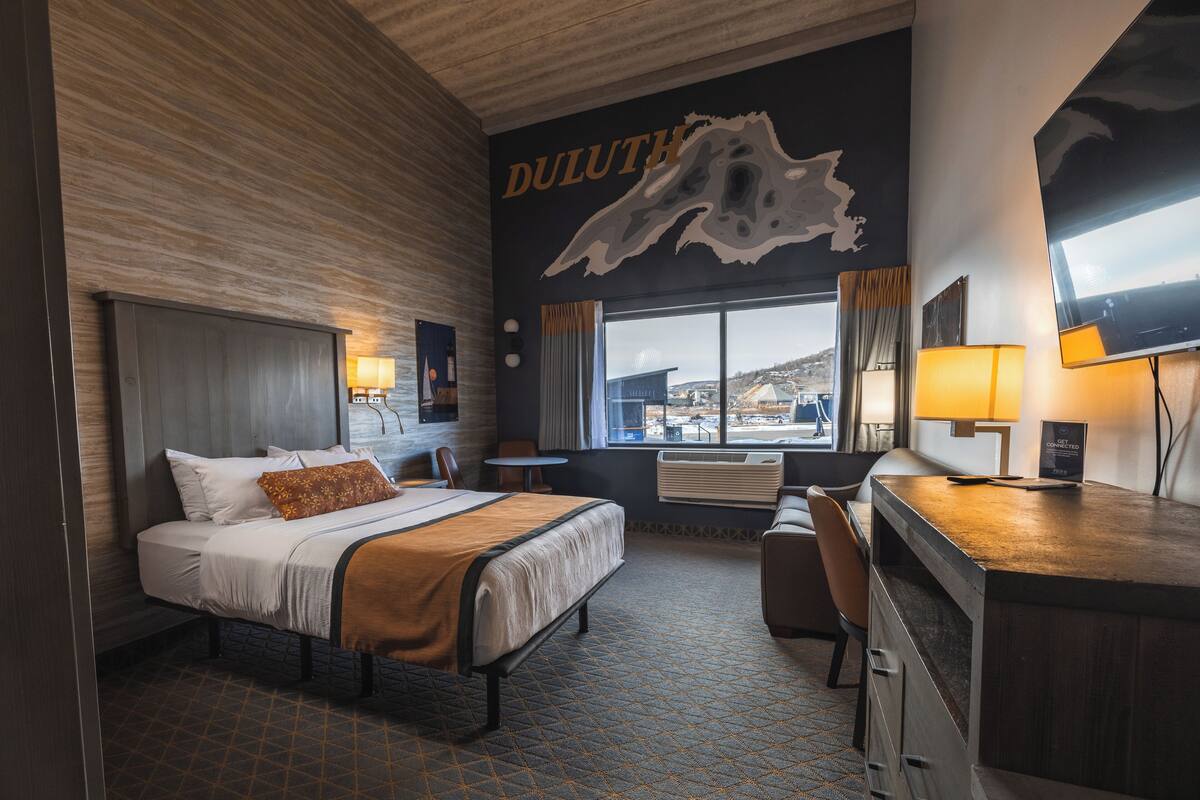 Une chambre d’hôtel avec un grand lit, un bureau, une chaise, une fenêtre donnant sur un beau paysage et un mur décoré d’une carte.