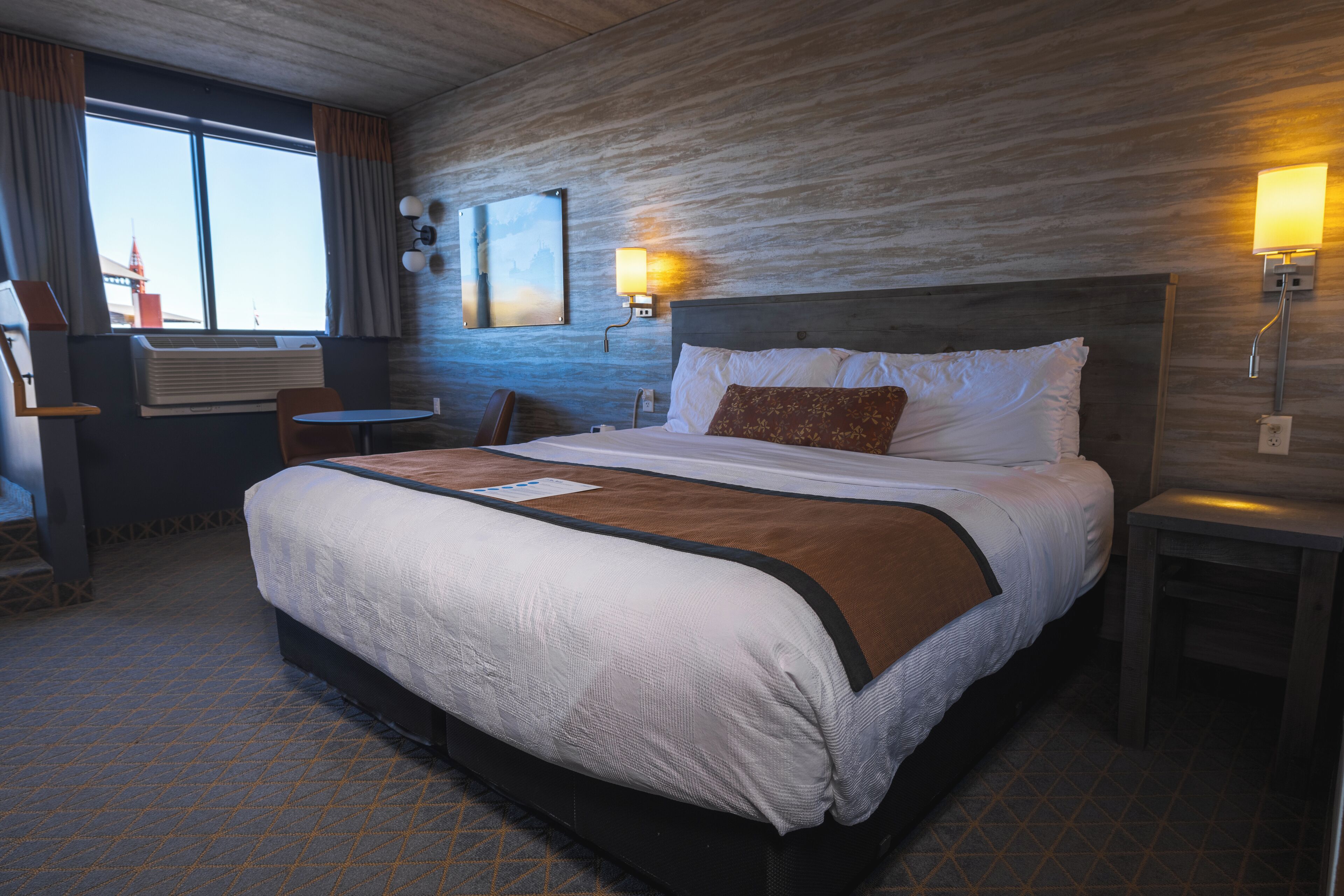 A hotel room with a large bed, a desk, and a chair. There is a window with curtains, a wall-mounted lamp, and an air conditioning unit.