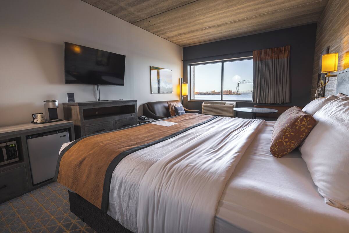 Une chambre d’hôtel avec un grand lit, un bureau avec une machine à café, un téléviseur fixé au mur et une fenêtre donnant sur un pont.