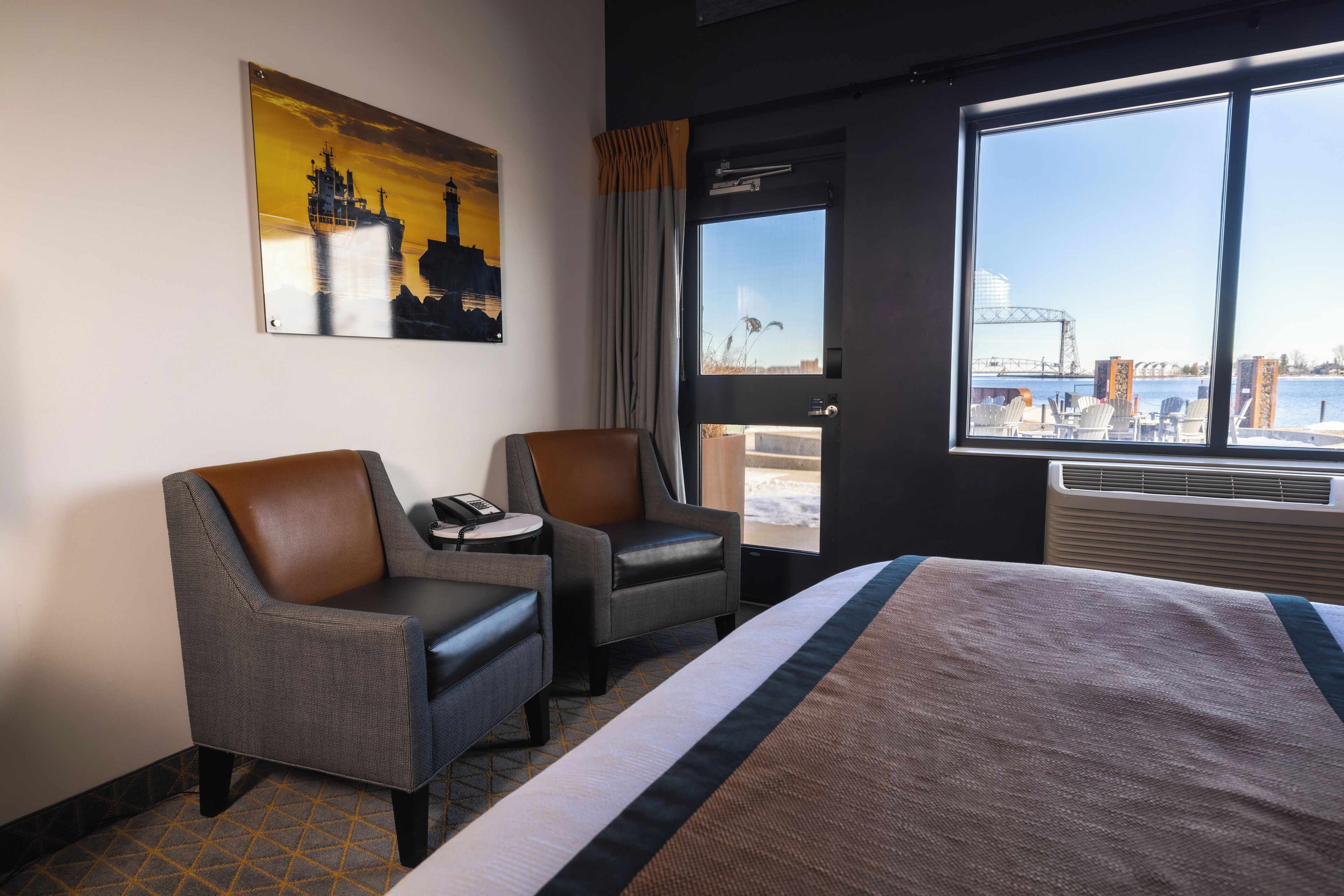 A hotel room with a large bed, two armchairs, a painting on the wall, and a view of the city through a large window.
