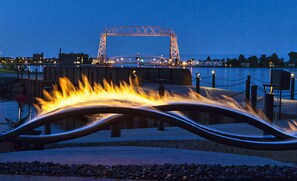 Un parque frente al agua con una escultura de metal, un puente y una masa de agua.