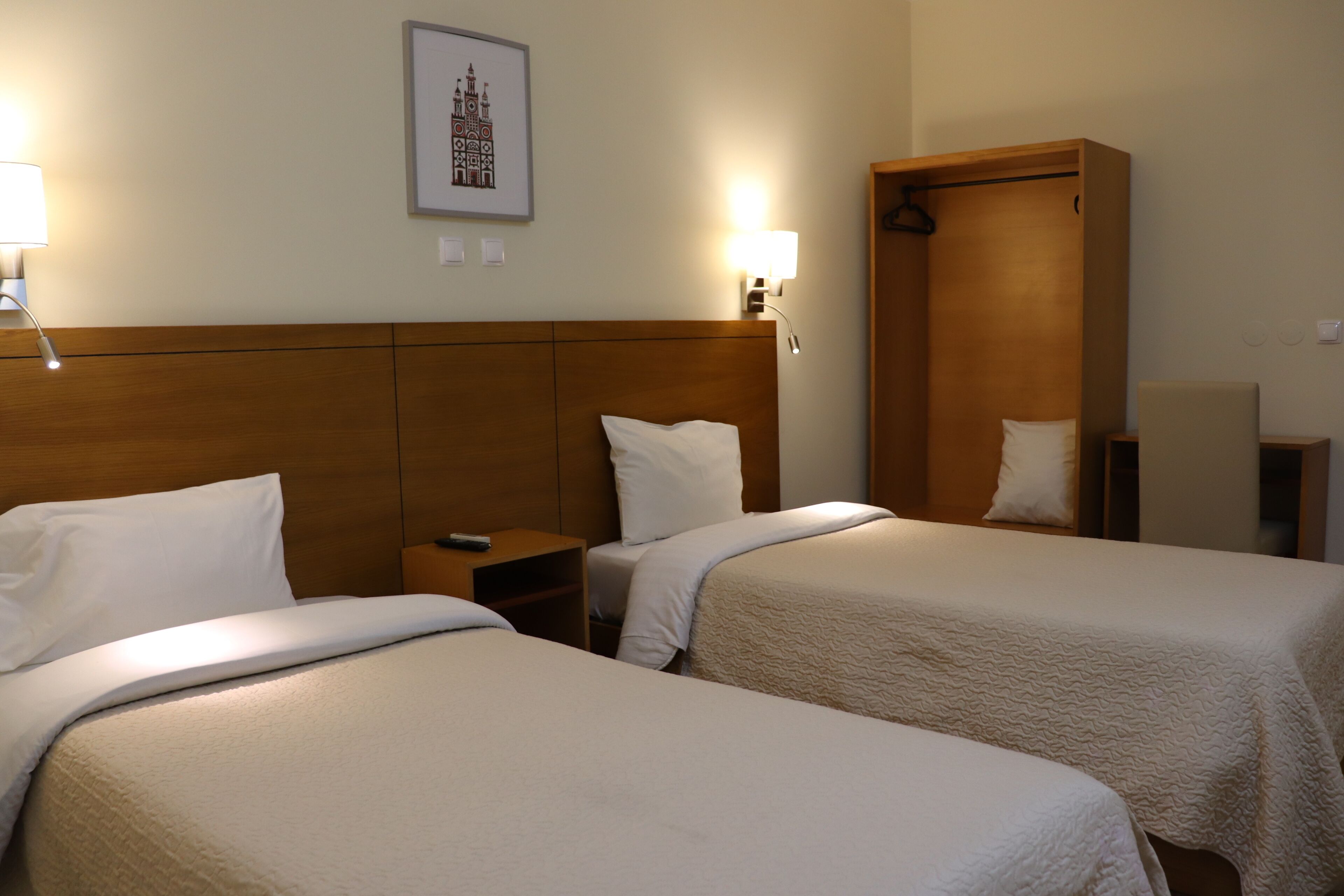 Een hotelkamer met twee bedden, een houten hoofdeinde, nachtlampjes, een lijst met een afbeelding aan de muur, en een houten garderobe.