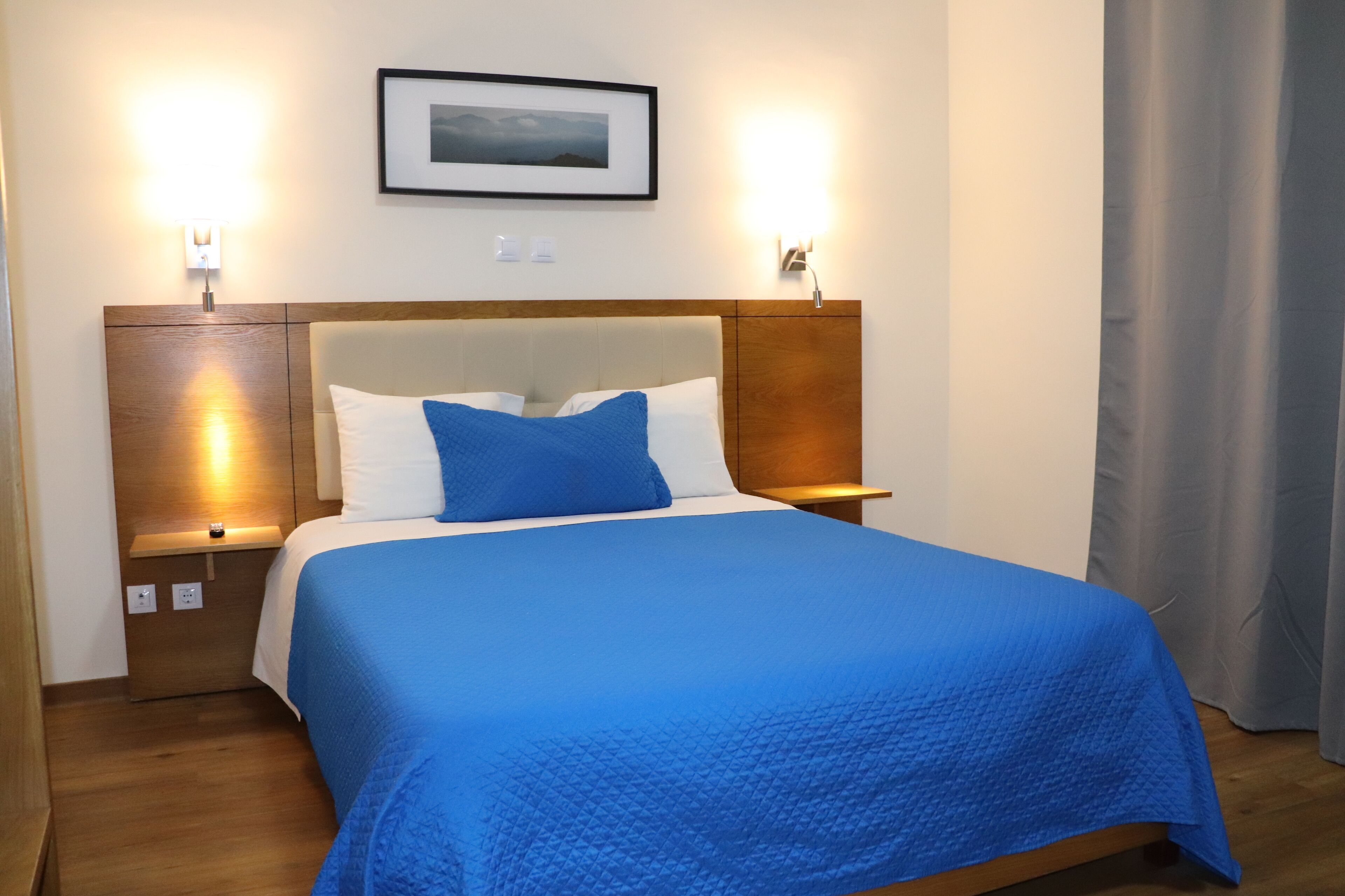 Een keurig opgemaakt bed met een blauwe sprei, twee bedlampen en een ingelijste foto aan de muur.