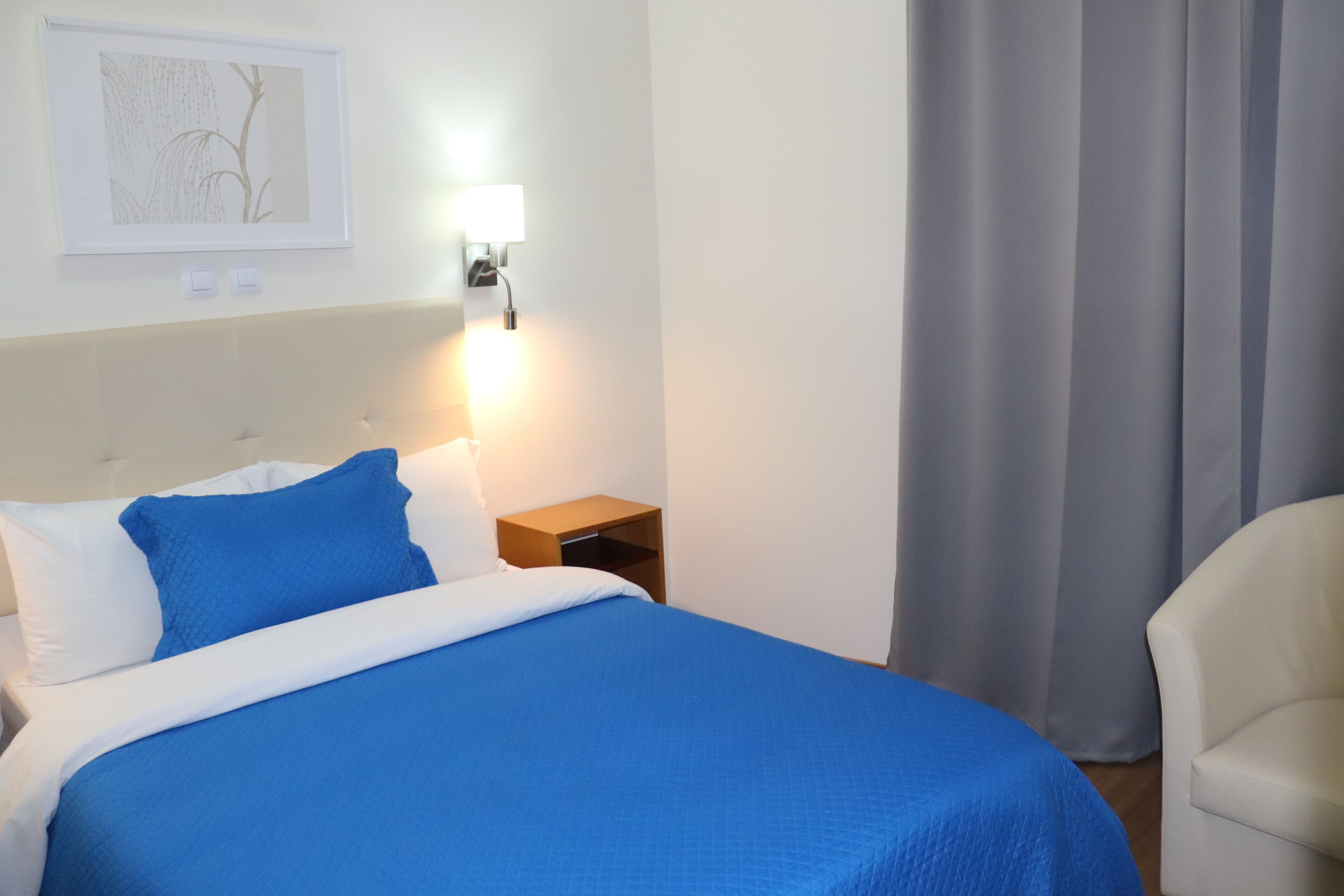 A neatly made bed with a blue bedspread, a bedside lamp, a framed picture on the wall, and a chair to the right.