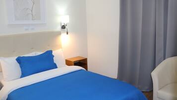 A neatly made bed with a blue bedspread, a bedside lamp, a framed picture on the wall, and a chair to the right.