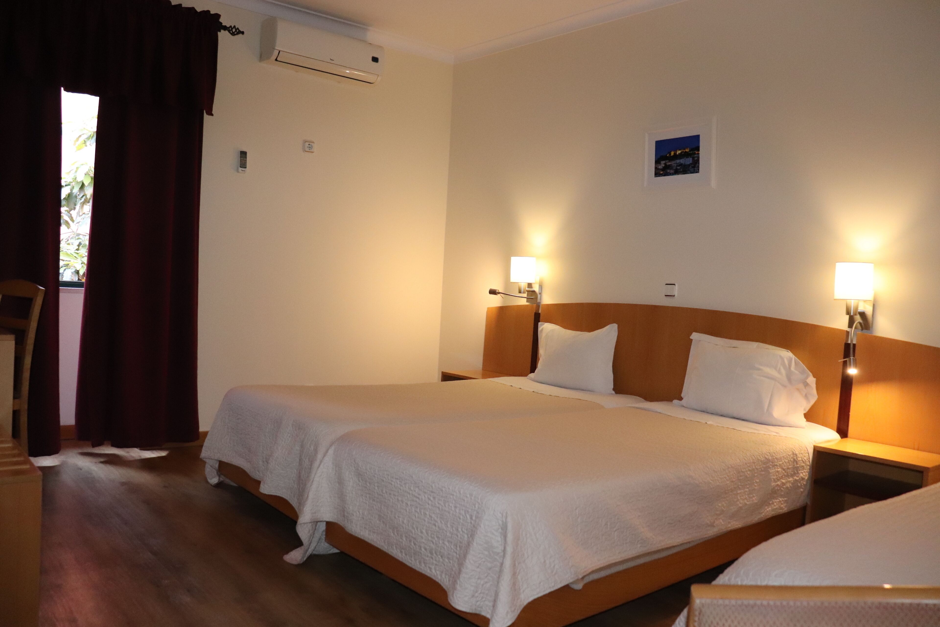 Habitación de hotel con dos camas, cabecera de madera, lámparas de noche, una ventana con cortinas y un equipo de aire acondicionado.