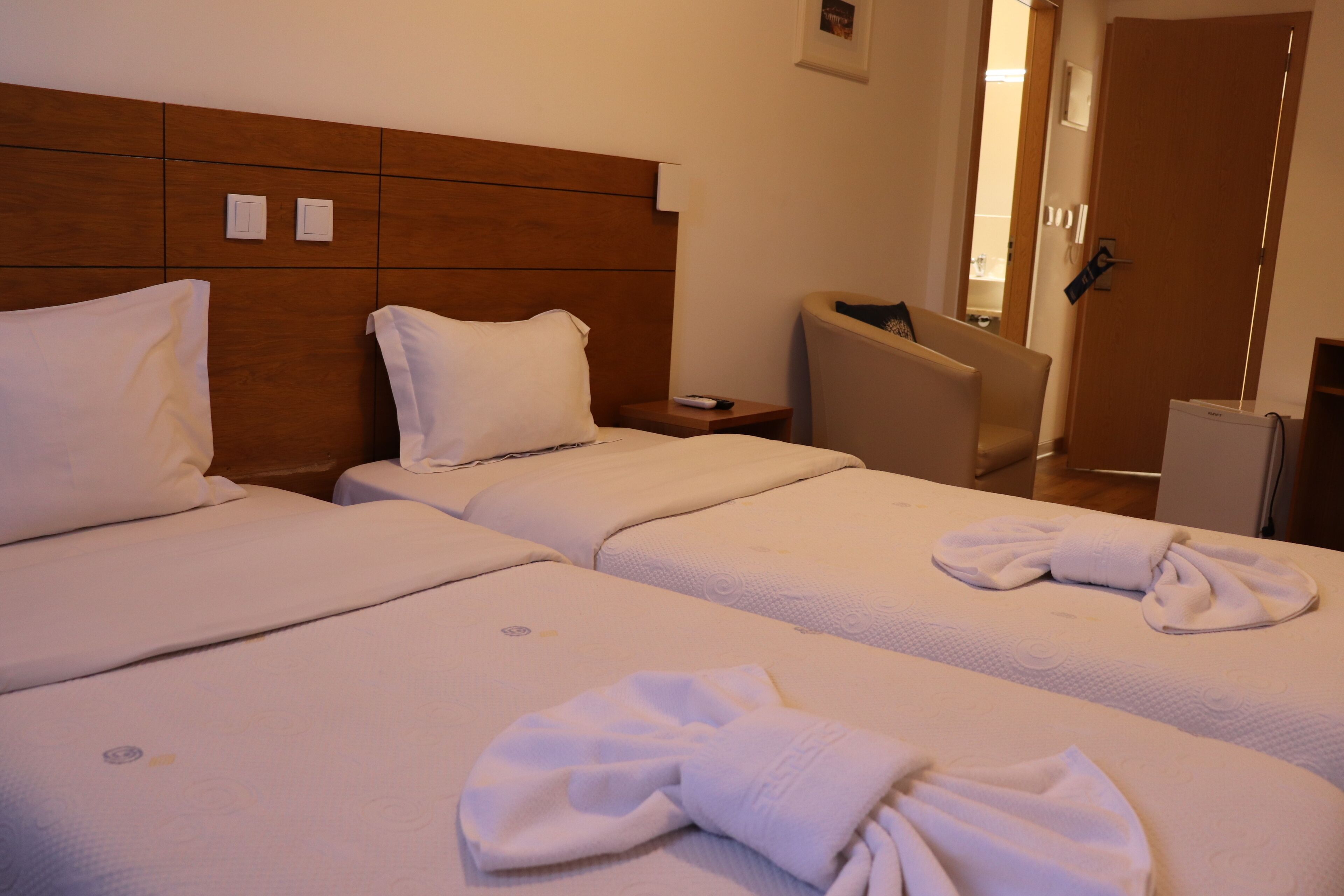A hotel room with two beds, a wooden headboard, a small bedside table with a remote control, a chair, and a door.
