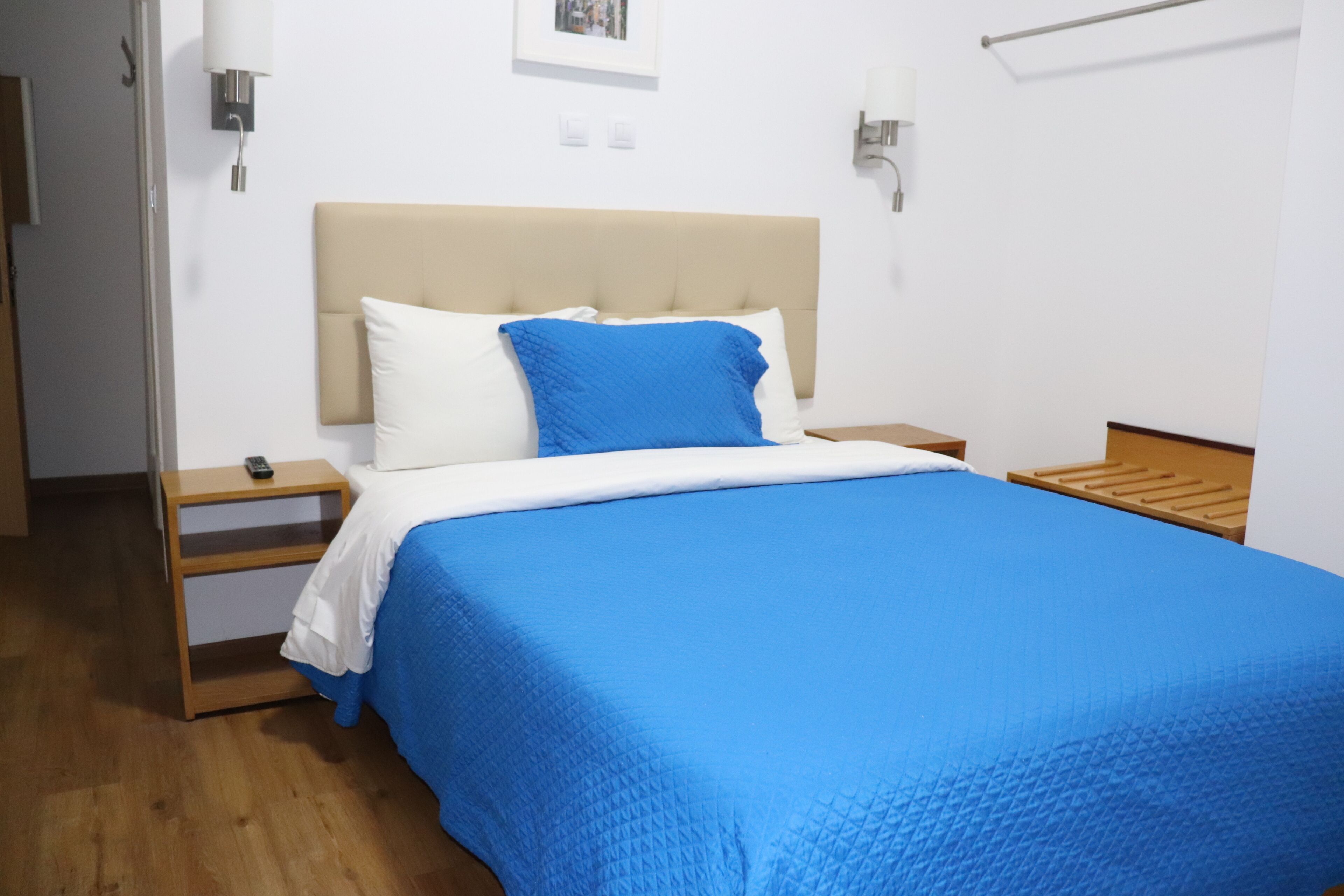Un lit bien fait, avec une couvre-lit bleue et des oreillers blancs, une table de chevet en bois avec une télécommande, et une tête de lit en bois.