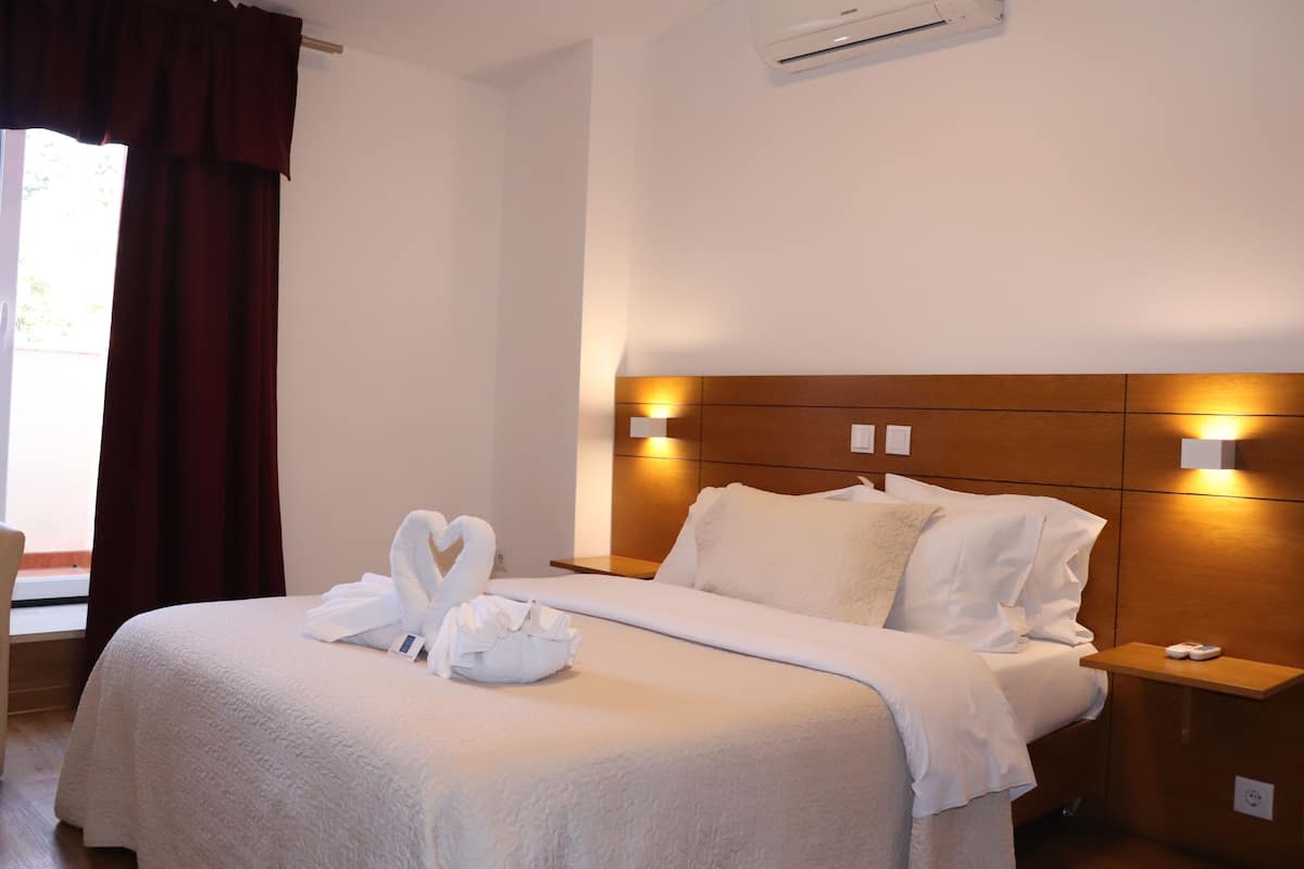 A hotel room with a neatly made bed, a wooden headboard, bedside lamps, and a window with curtains.