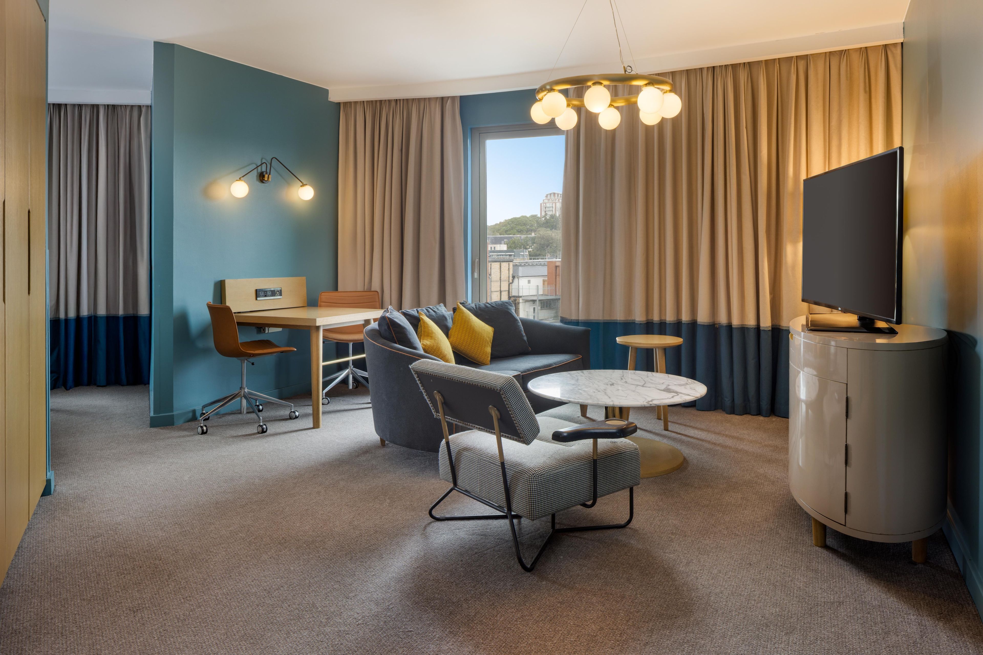 Ein modernes Hotelzimmer mit einer Couch, Sesseln, einem Esstisch, einem Fernseher und einem großen Fenster mit Vorhängen.