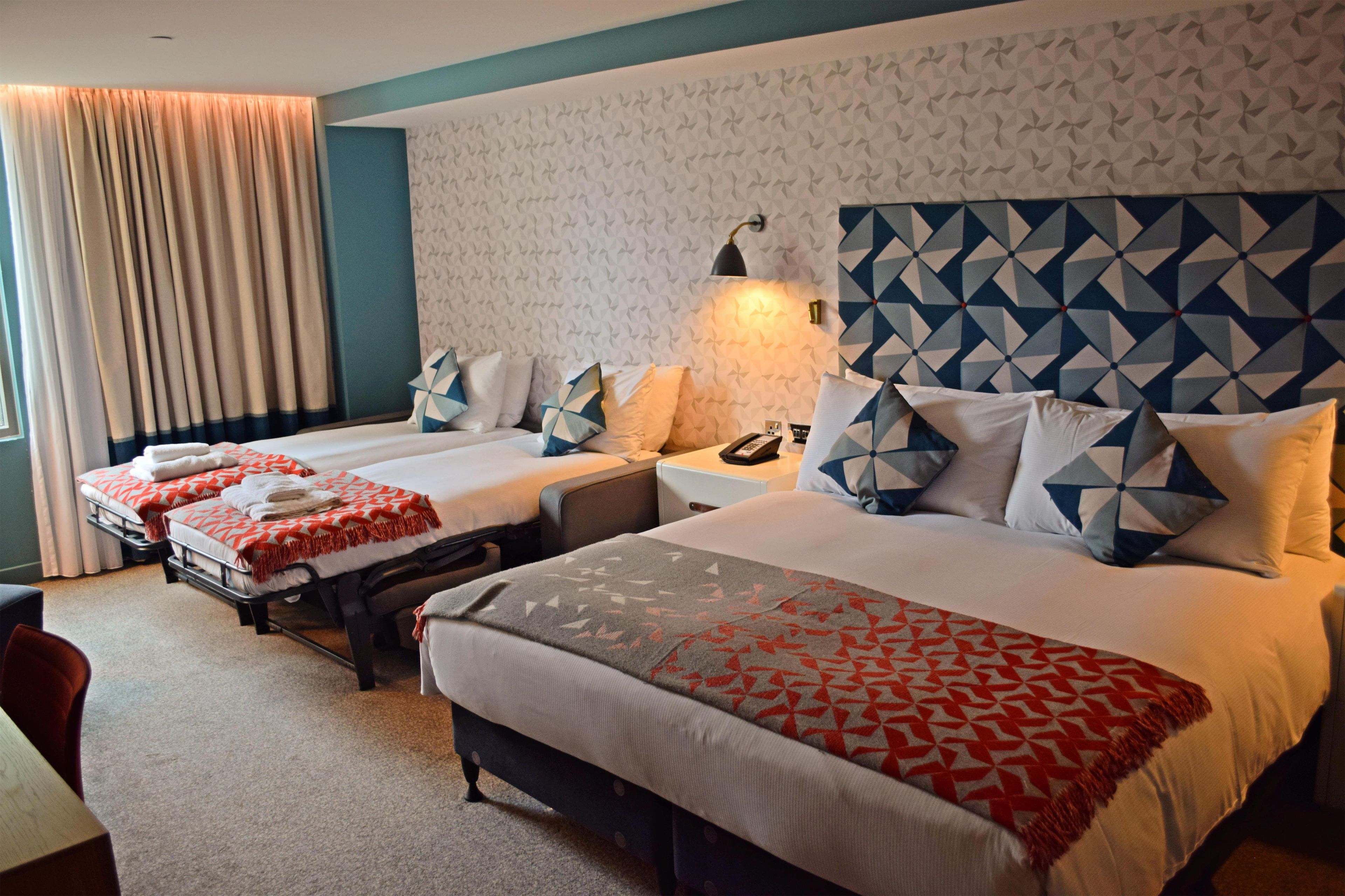A hotel room with two beds, a desk, a chair, a lamp, and a patterned wall.
