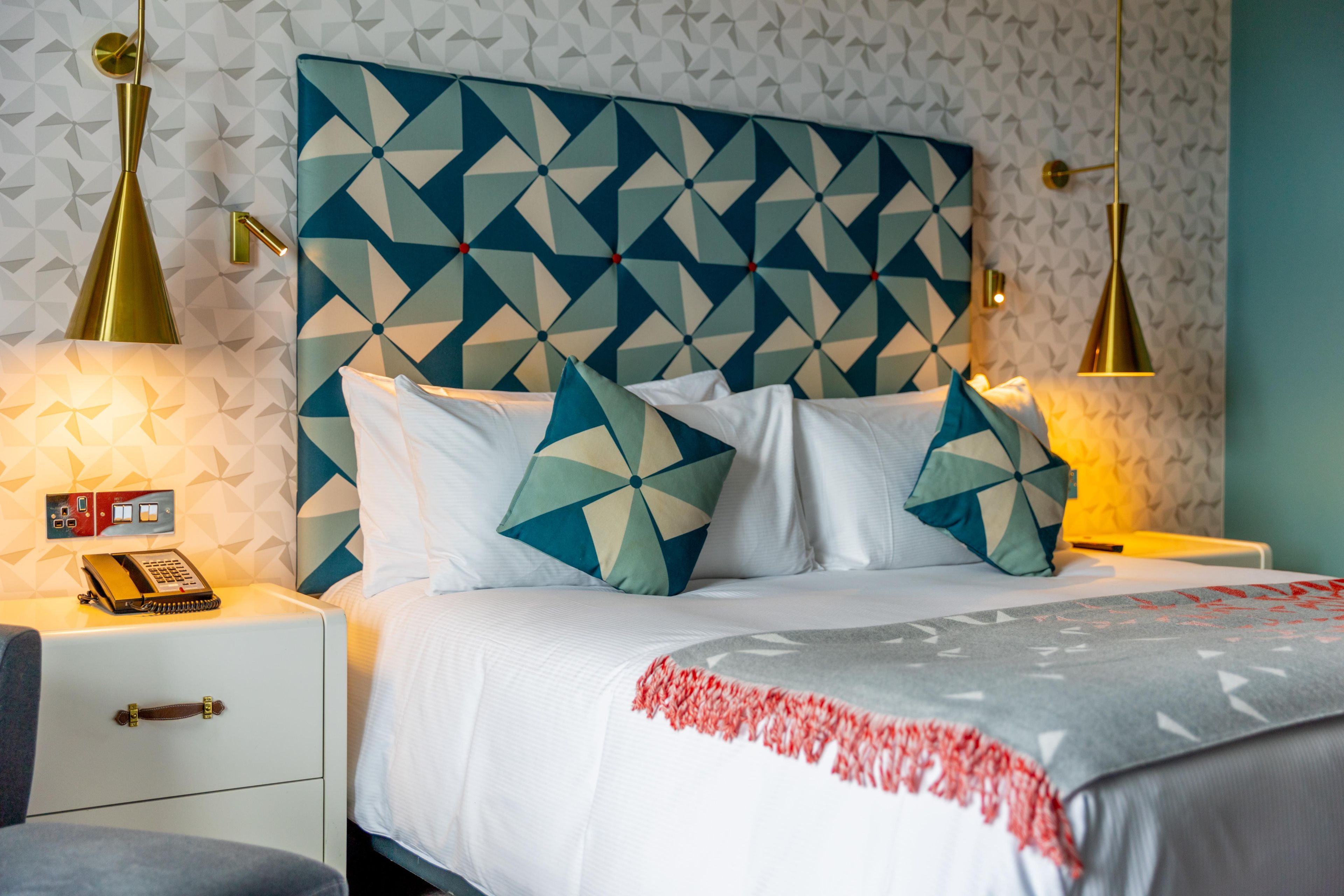 Una habitación de hotel con cabecera de diseño geométrico, una cama con ropa de cama blanca y cojines decorativos, lámparas de noche, un teléfono y un pequeño escritorio con lámpara.