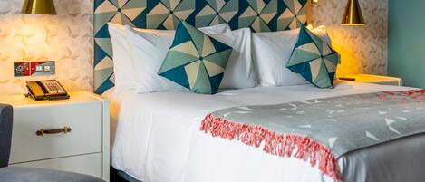 Una habitación de hotel con cabecera de diseño geométrico, una cama con ropa de cama blanca y cojines decorativos, lámparas de noche, un teléfono y un pequeño escritorio con lámpara.