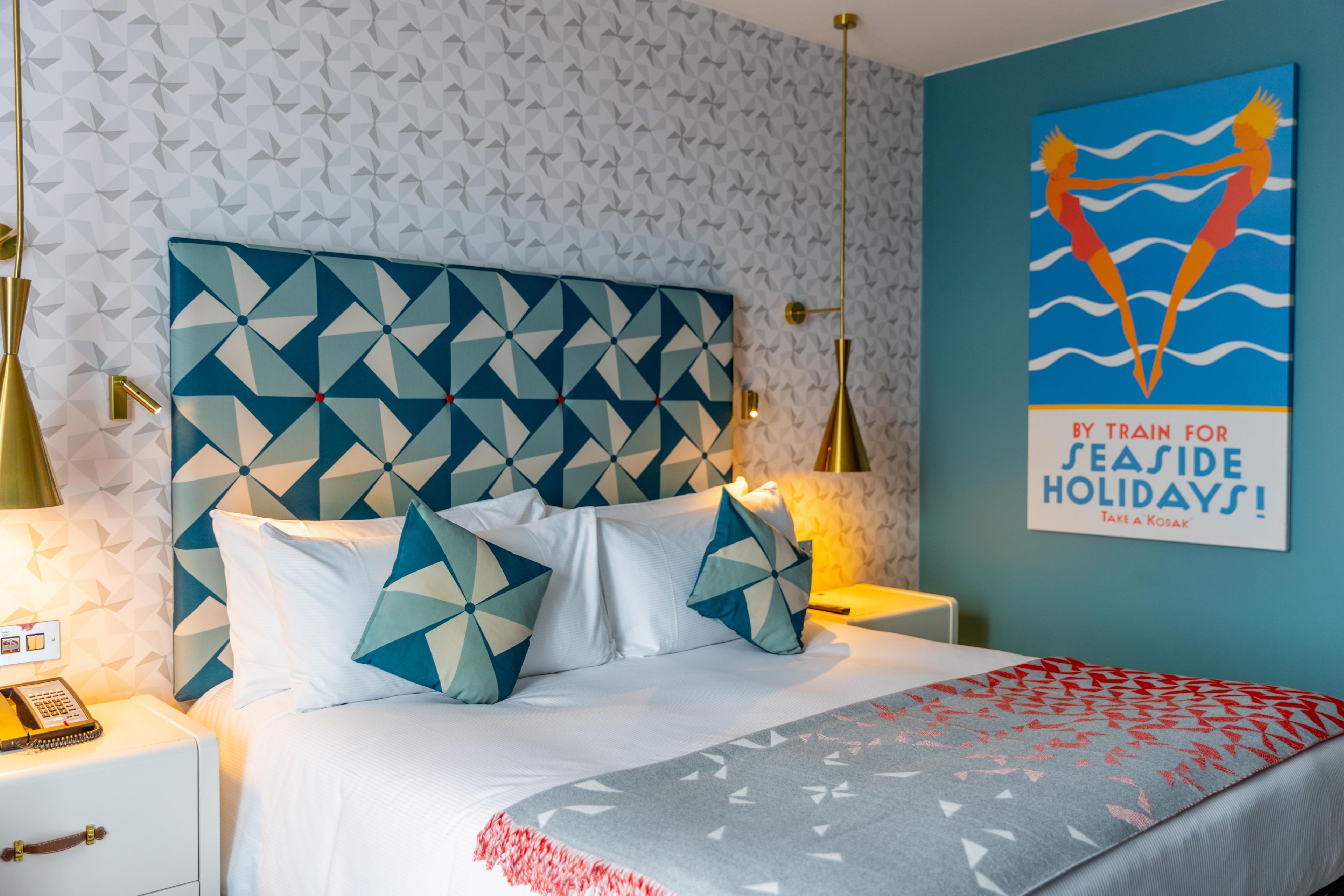 A hotel room with a geometric-patterned headboard, a bed with white bedding, a blue and white throw blanket, and a wall with a geometric wallpaper and a poster.