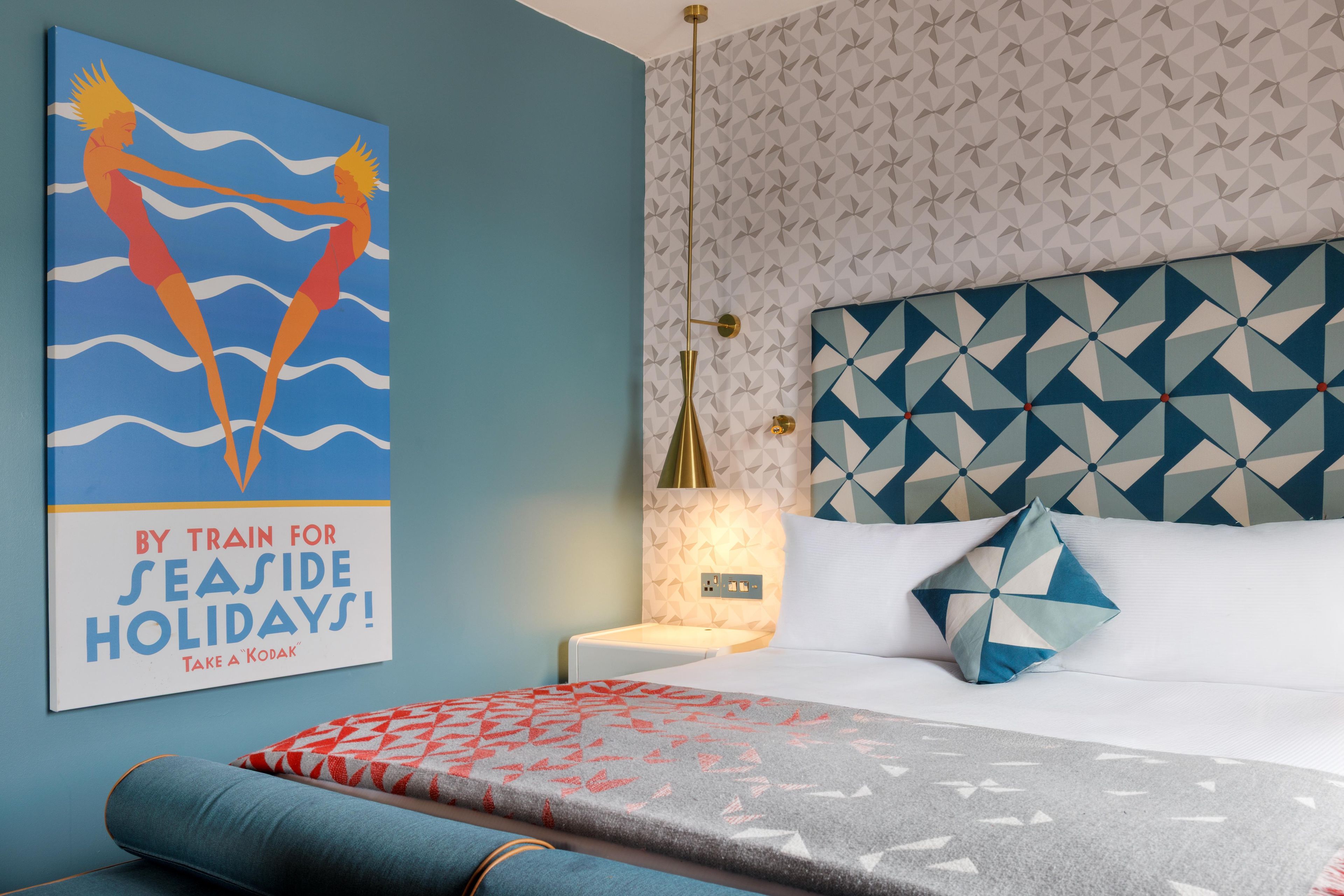 A hotel room with a geometric patterned headboard, a bed with a patterned throw, a nightstand with a lamp, and a poster on the wall.